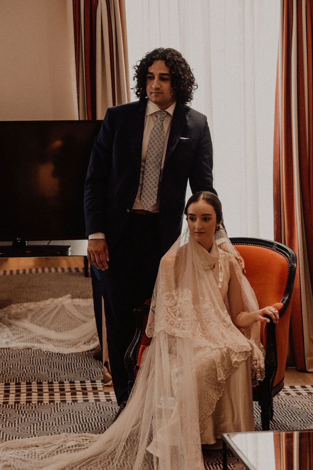 Bride in lace veil seated in orange chair with groom in navy suit standing beside her in elegant hotel room.