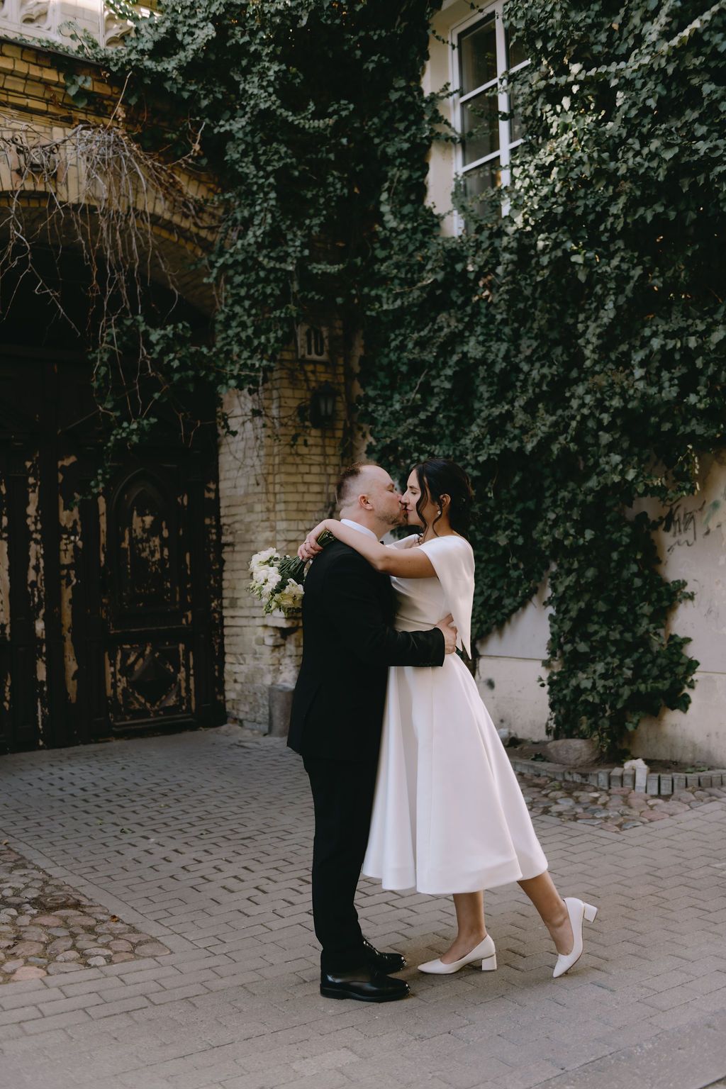 Bride and groom share a romantic kiss outdoors, surrounded by ivy-covered walls and ornate gates.