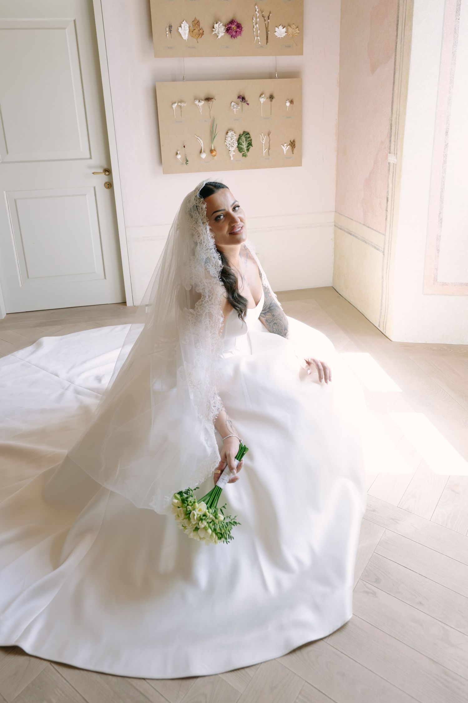 Smiling bride in a voluminous satin wedding gown and veil holds a bouquet while seated on a light wooden floor.