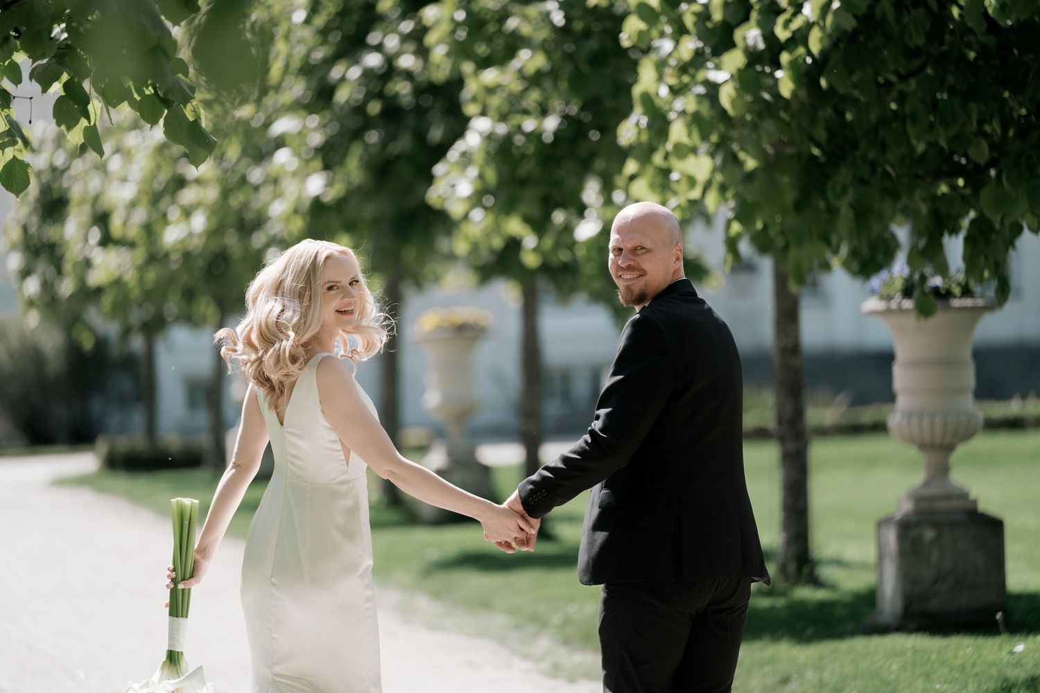 Smiling bride in a white dress holds hands with groom in a dark suit while walking along a sunlit garden path lined with trees.