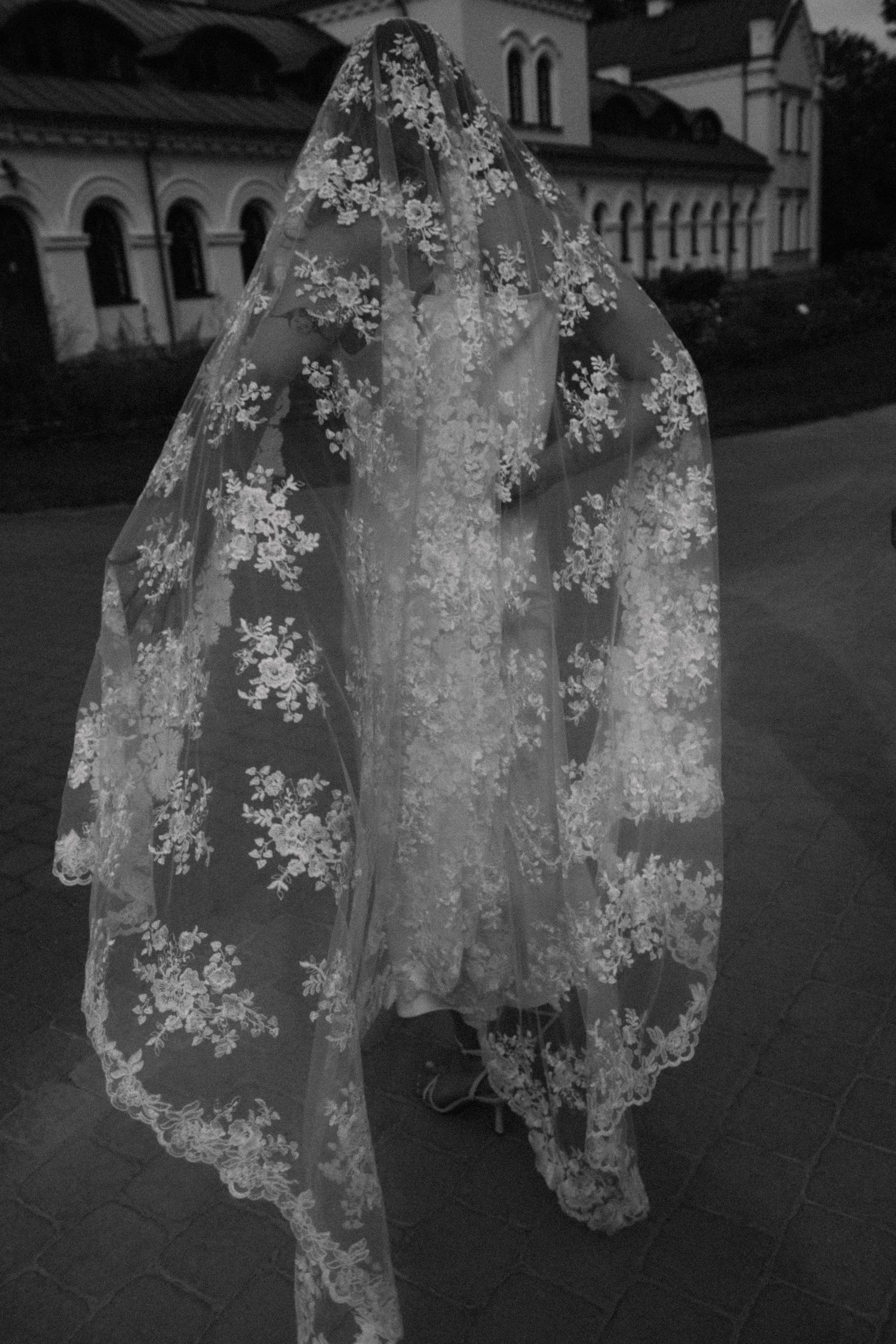 Bride covered head to toe in a long ivory lace wedding veil with floral embroidery, standing on a paved courtyard in black and white.