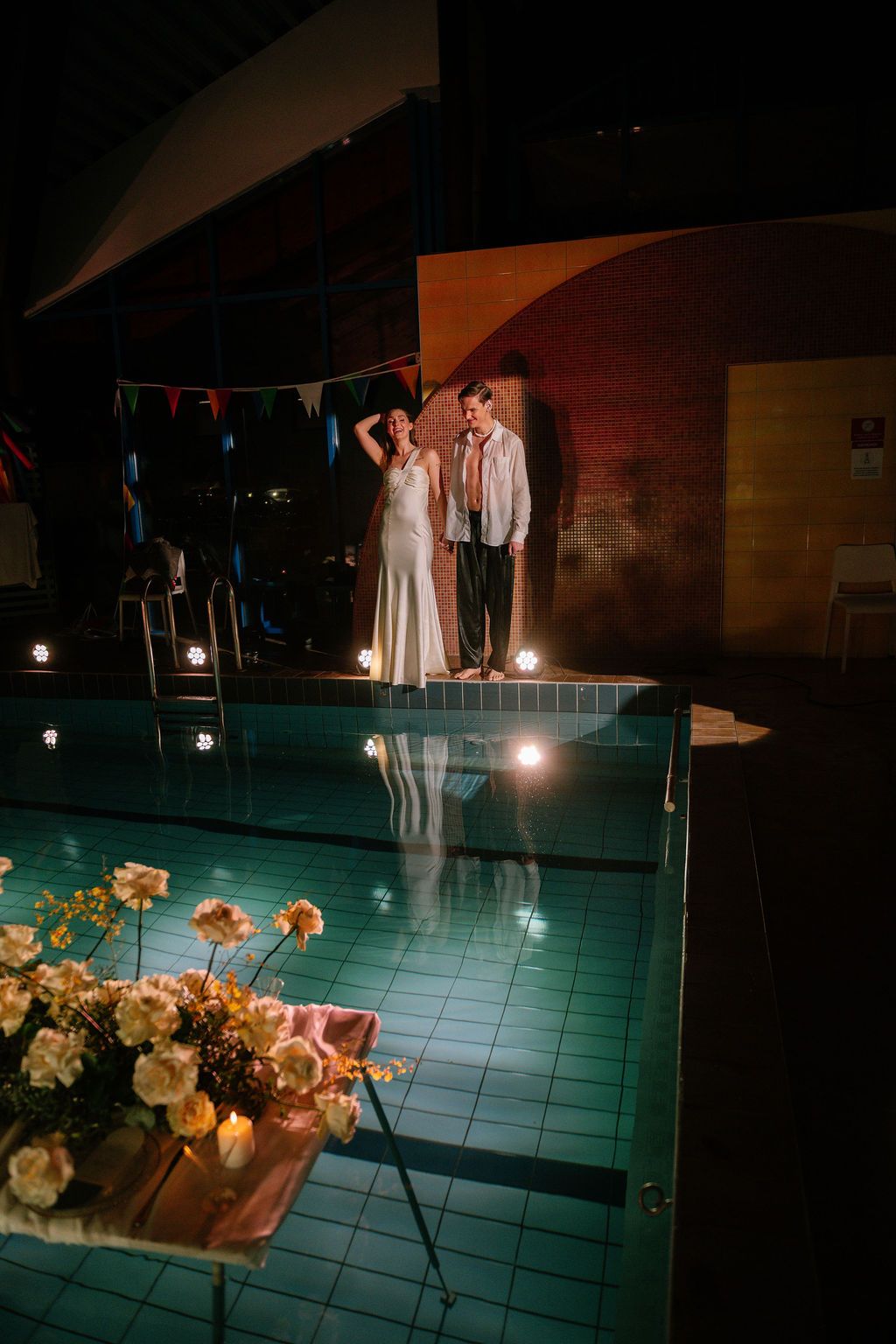 Bride in a white gown and groom in a vest stand at the edge of an indoor pool at night, with floating white roses and candles.