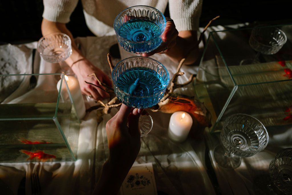 Hands holding crystal glasses filled with blue drinks beside a lit white candle on a dark table, evoking a mystical ritual atmosphere.