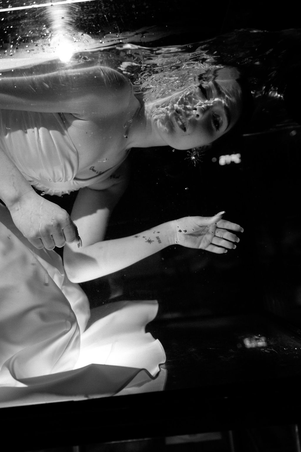Black and white underwater portrait of a woman in a flowing white gown floating gracefully with sunlight shimmering at the water's surface.