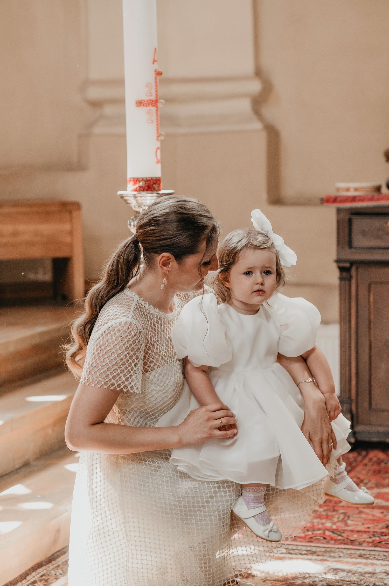 A parent and child in matching white outfits share tender moments in a sunlit church setting.