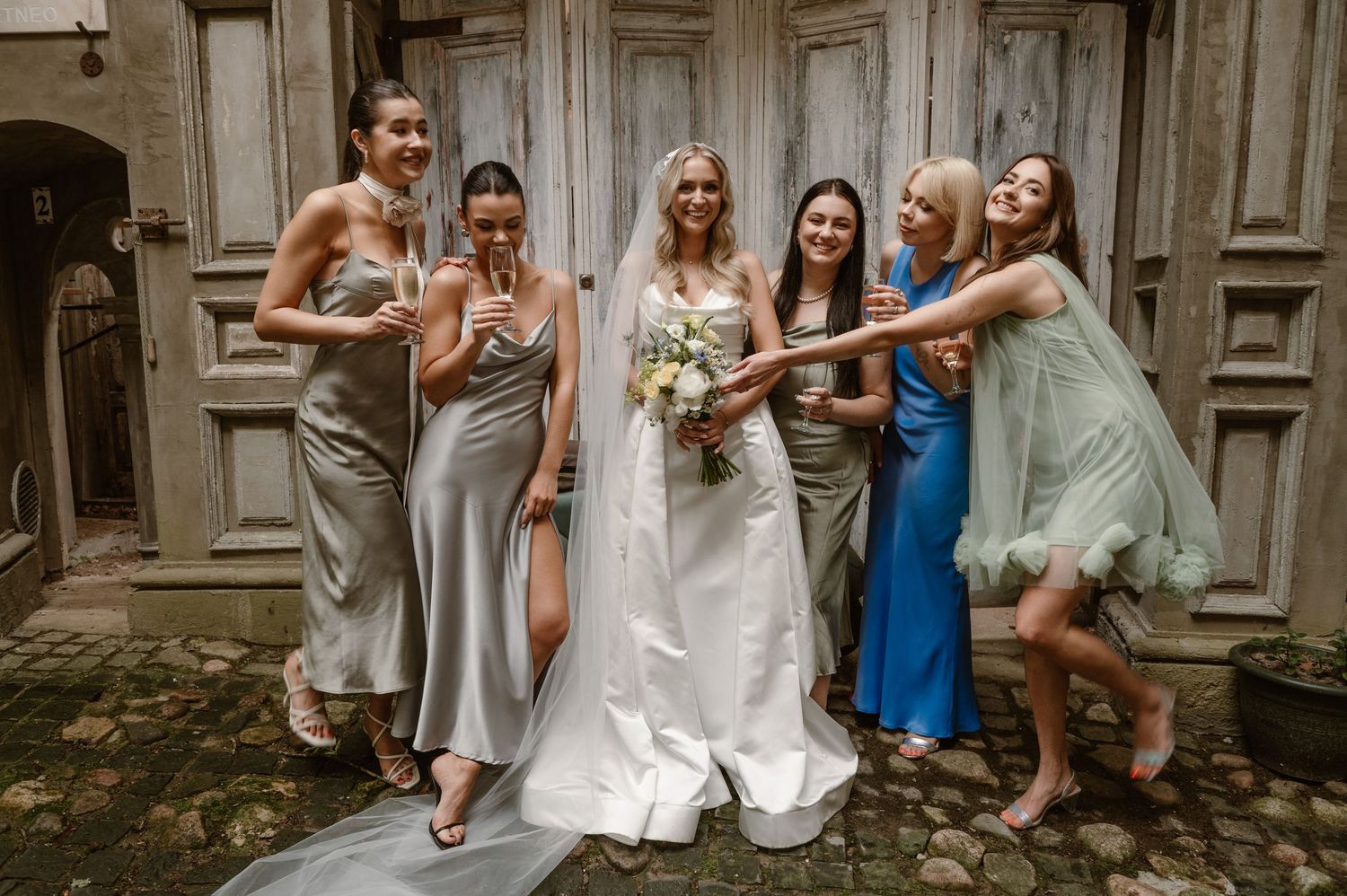 A wedding party in elegant dresses poses playfully against rustic wooden doors at a historic venue.