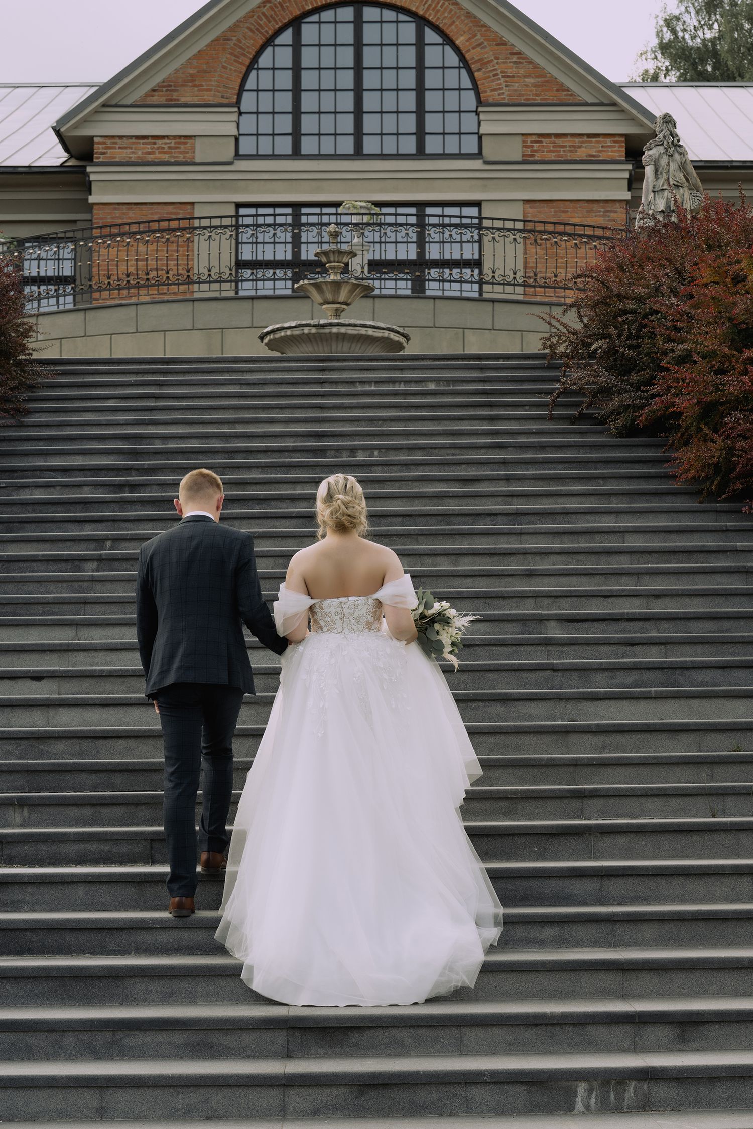 A couple in wedding attire ascending stone steps in front of a historic brick building with arched windows.