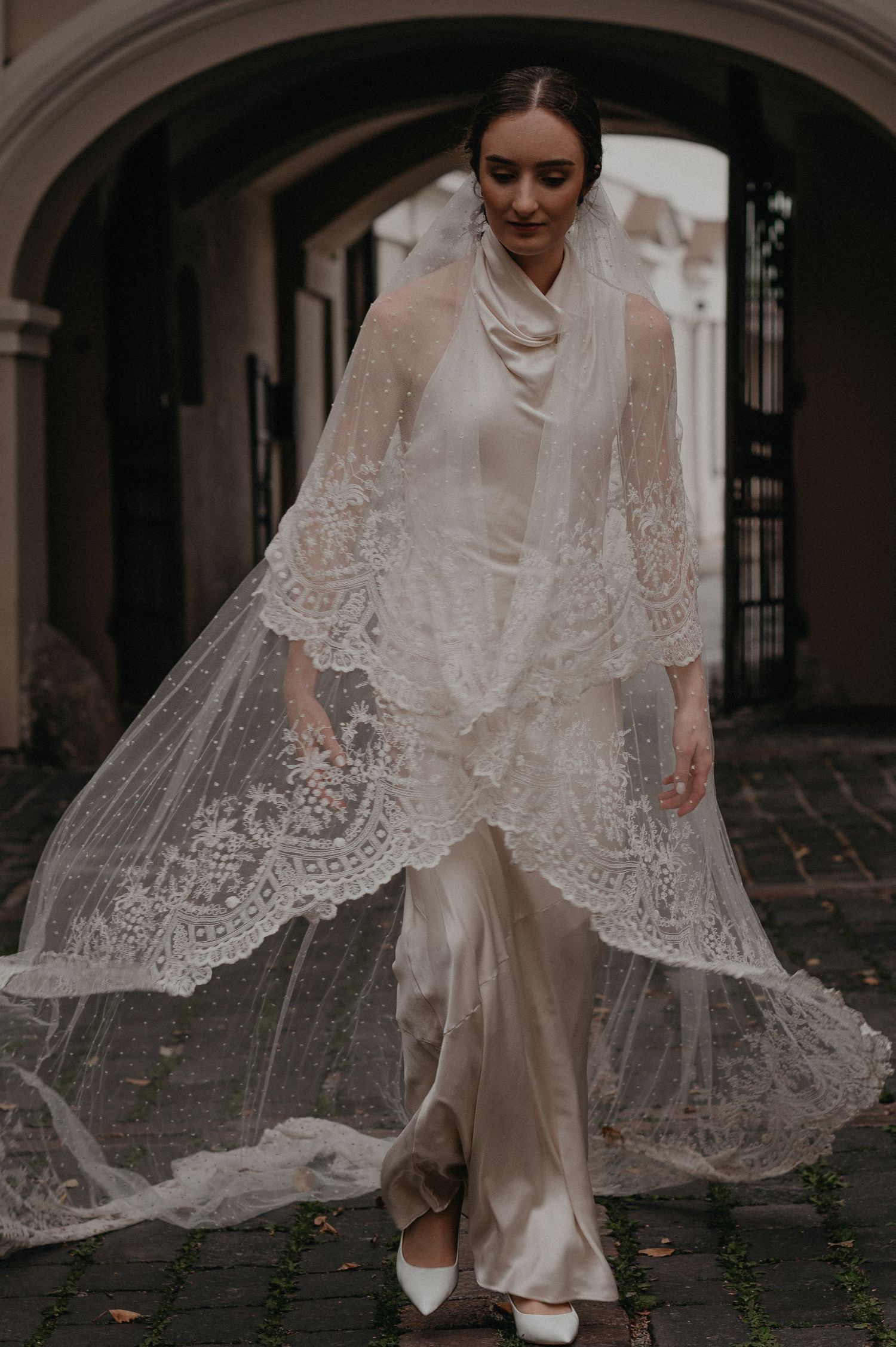 Bride in vintage-inspired white lace wedding dress and veil twirling in an arched courtyard.