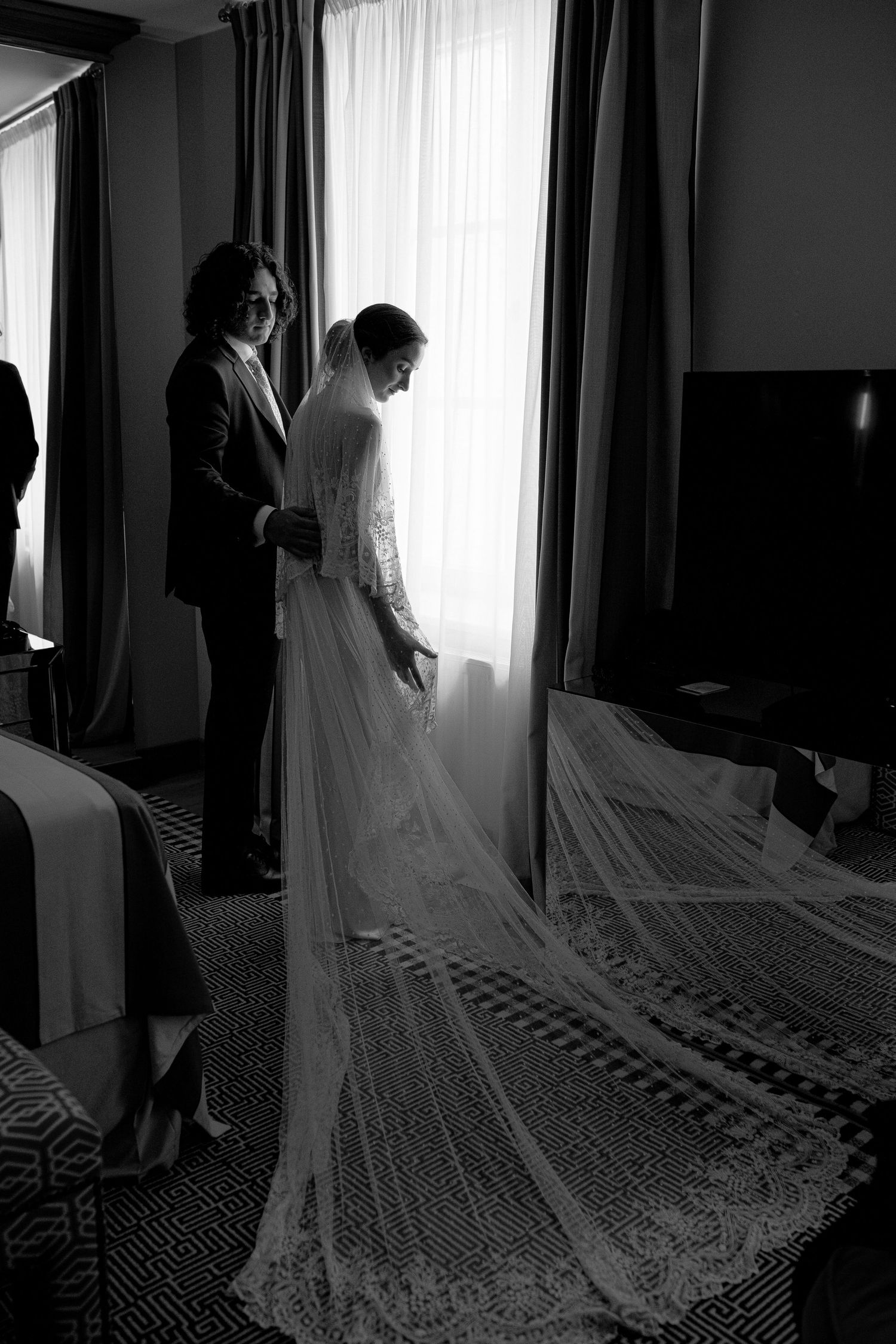 A black and white photo showing a wedding dress with an elegant lace train by a window in a hotel room.