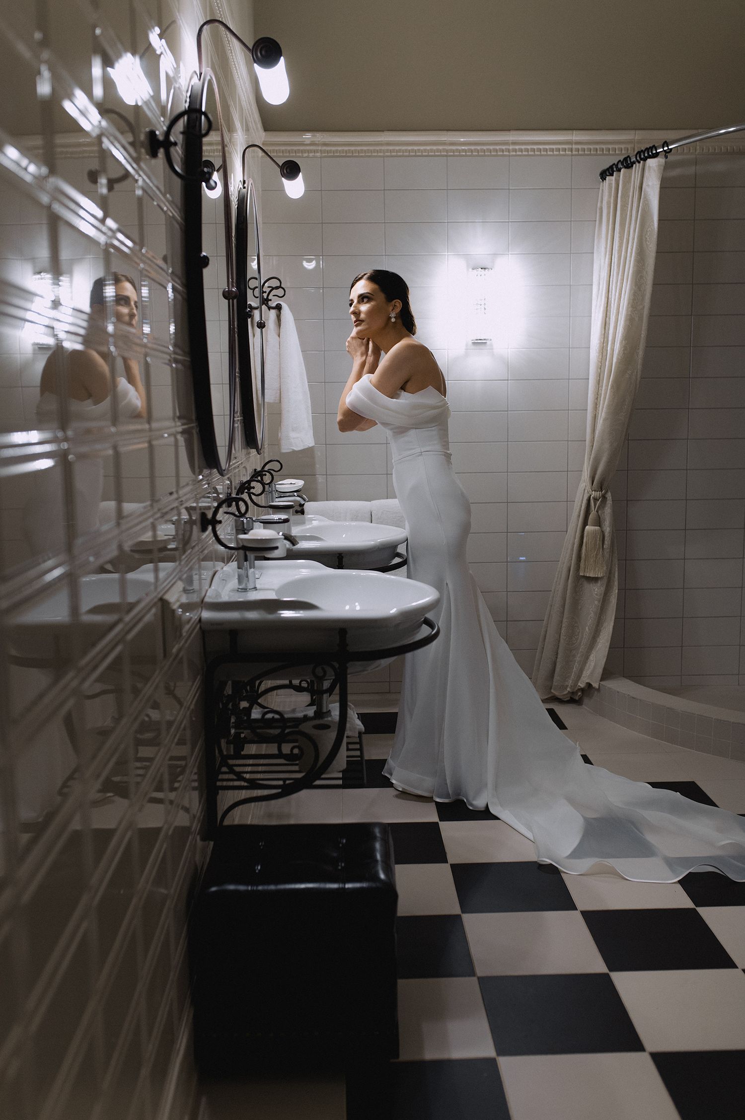 A person in a long white dress stands in an elegant black and white checkered bathroom with vintage pedestal sinks.