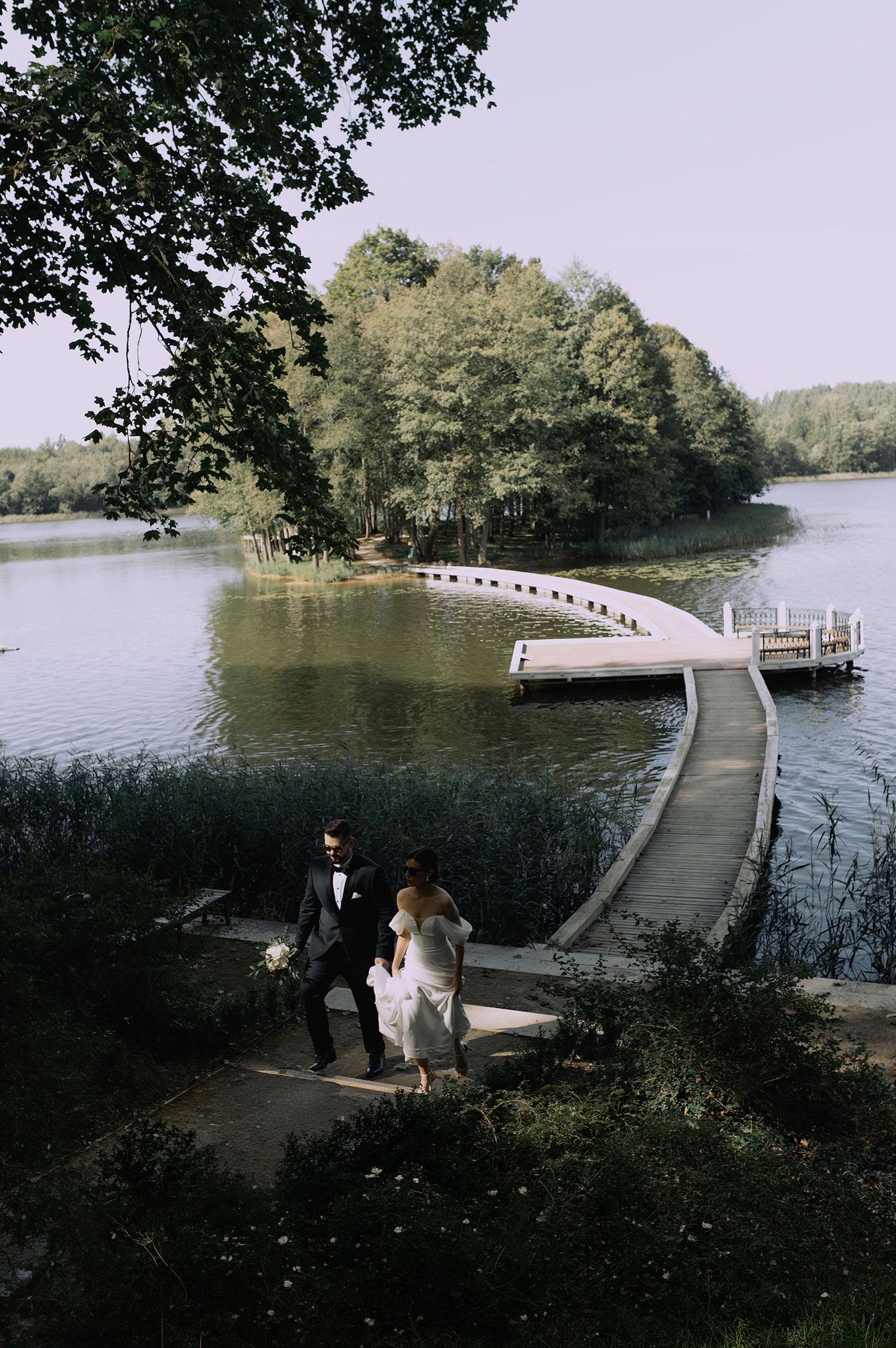 Wooden dock pathway extends over tranquil lake waters surrounded by lush green trees on a peaceful summer day.