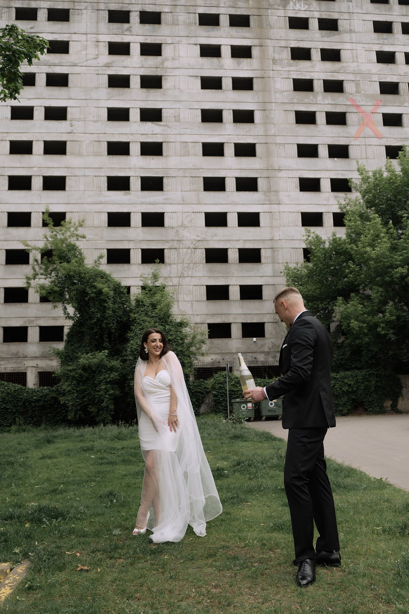 A couple shares a wedding moment in front of an industrial concrete building with trees in the foreground.