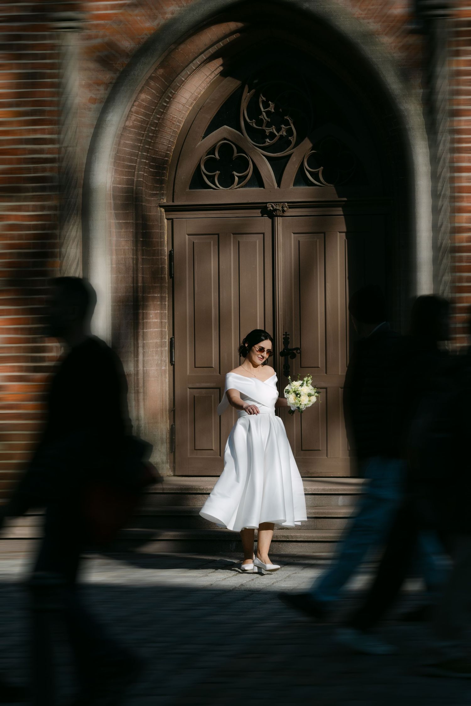Bride in white midi dress holding bouquet stands before ornate church doors as silhouetted figures pass by.