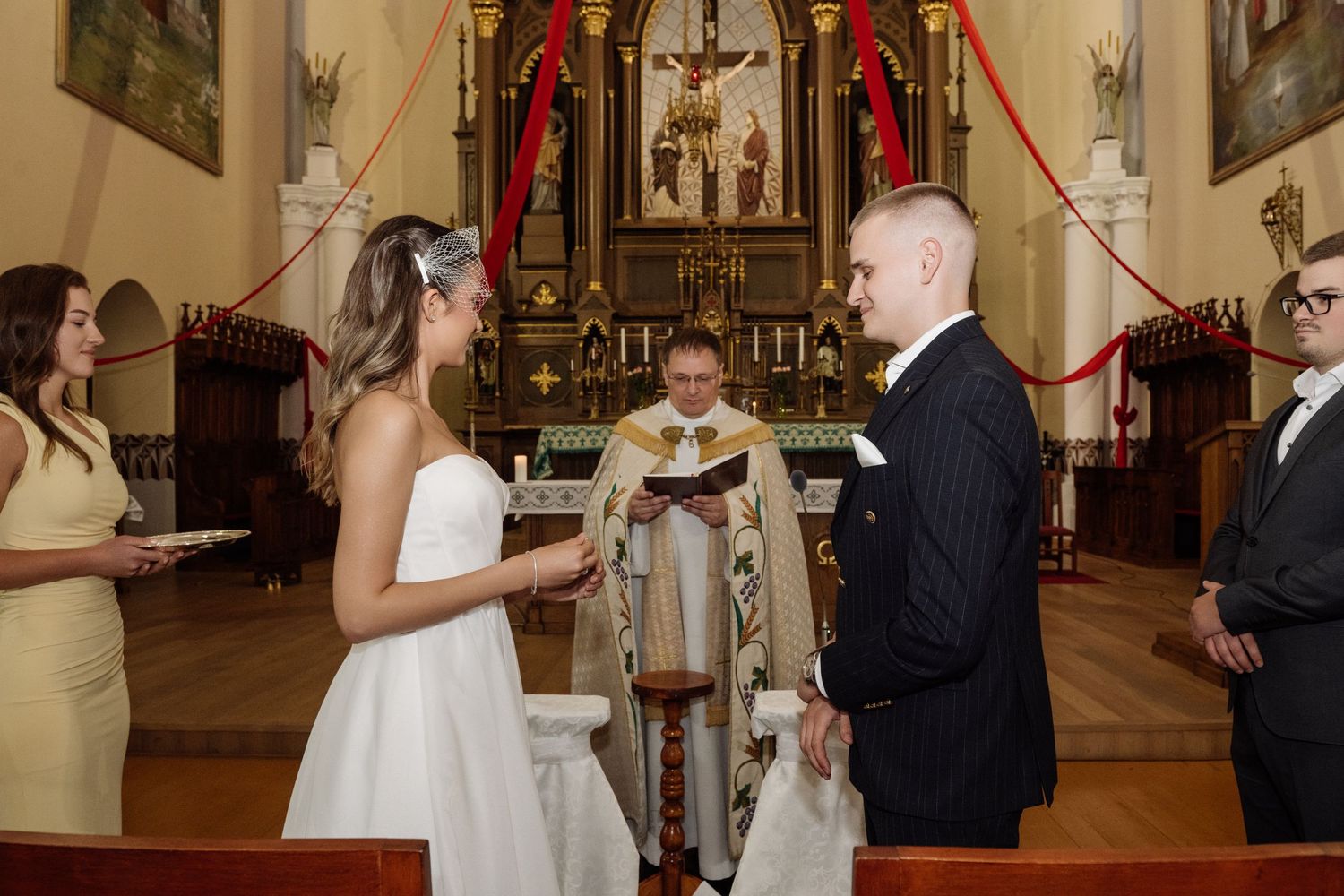 Bride in a white gown and groom in a dark suit exchange vows before a priest at the altar during a church wedding ceremony.