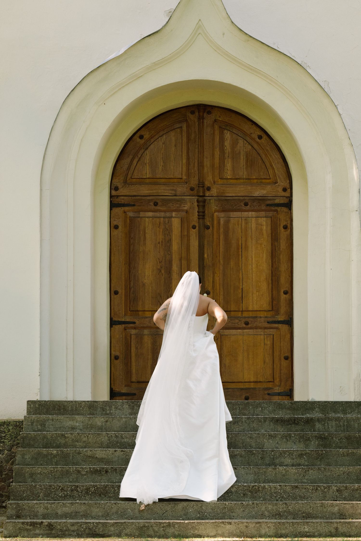 A bride in a long white gown and veil climbs stone steps toward a large arched wooden church door.