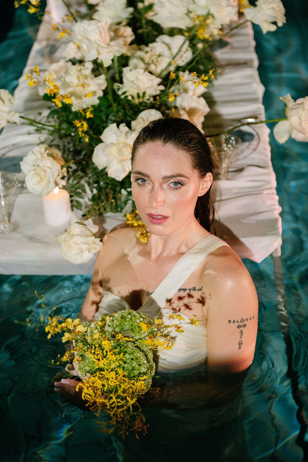 Bride in a white gown stands waist-deep in a pool holding a yellow floral bouquet beside a floating table of white roses and candles.