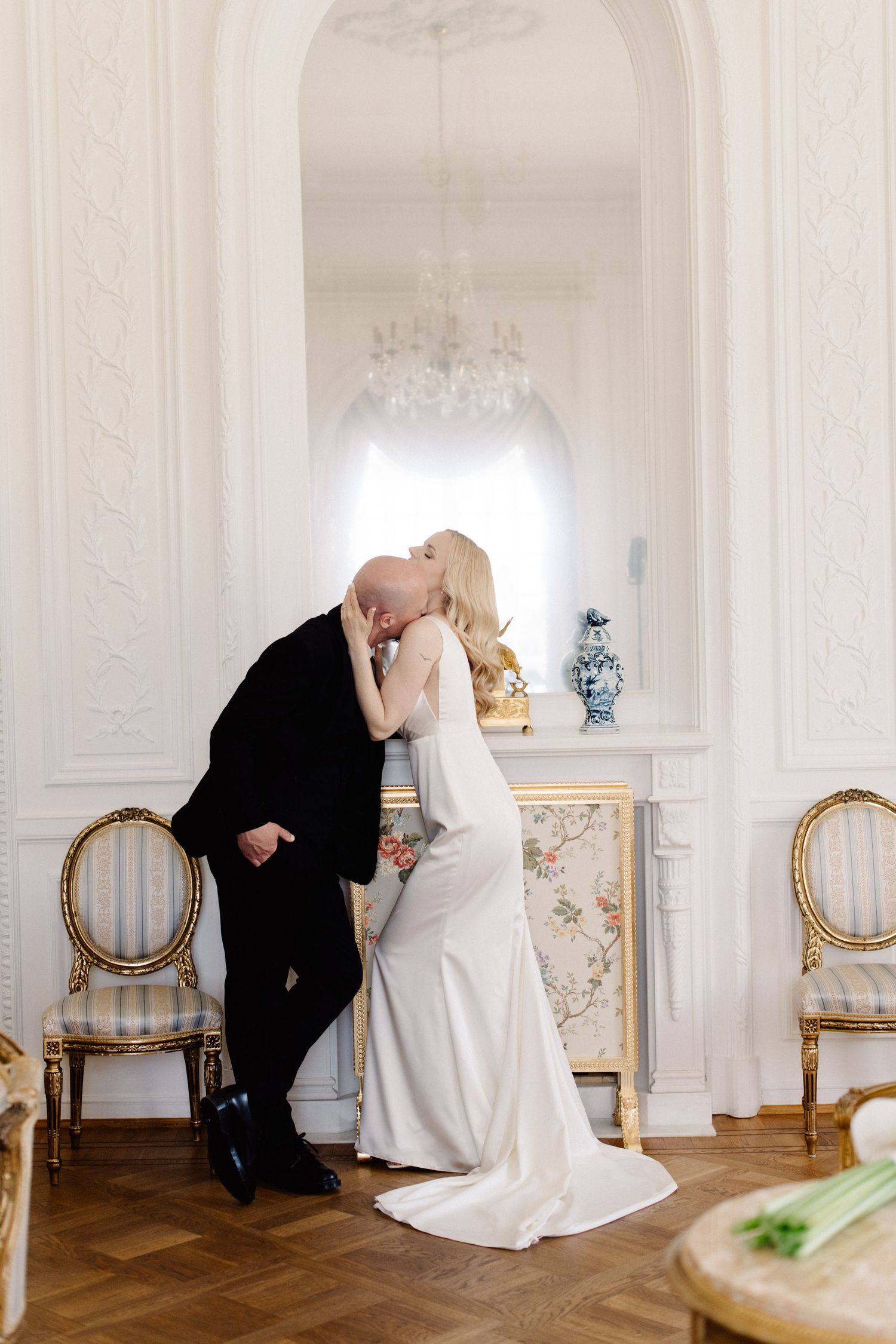 Bride in a white satin wedding dress kisses a groom in a black suit in a luxurious room with a chandelier, arched mirror and gilded chairs.