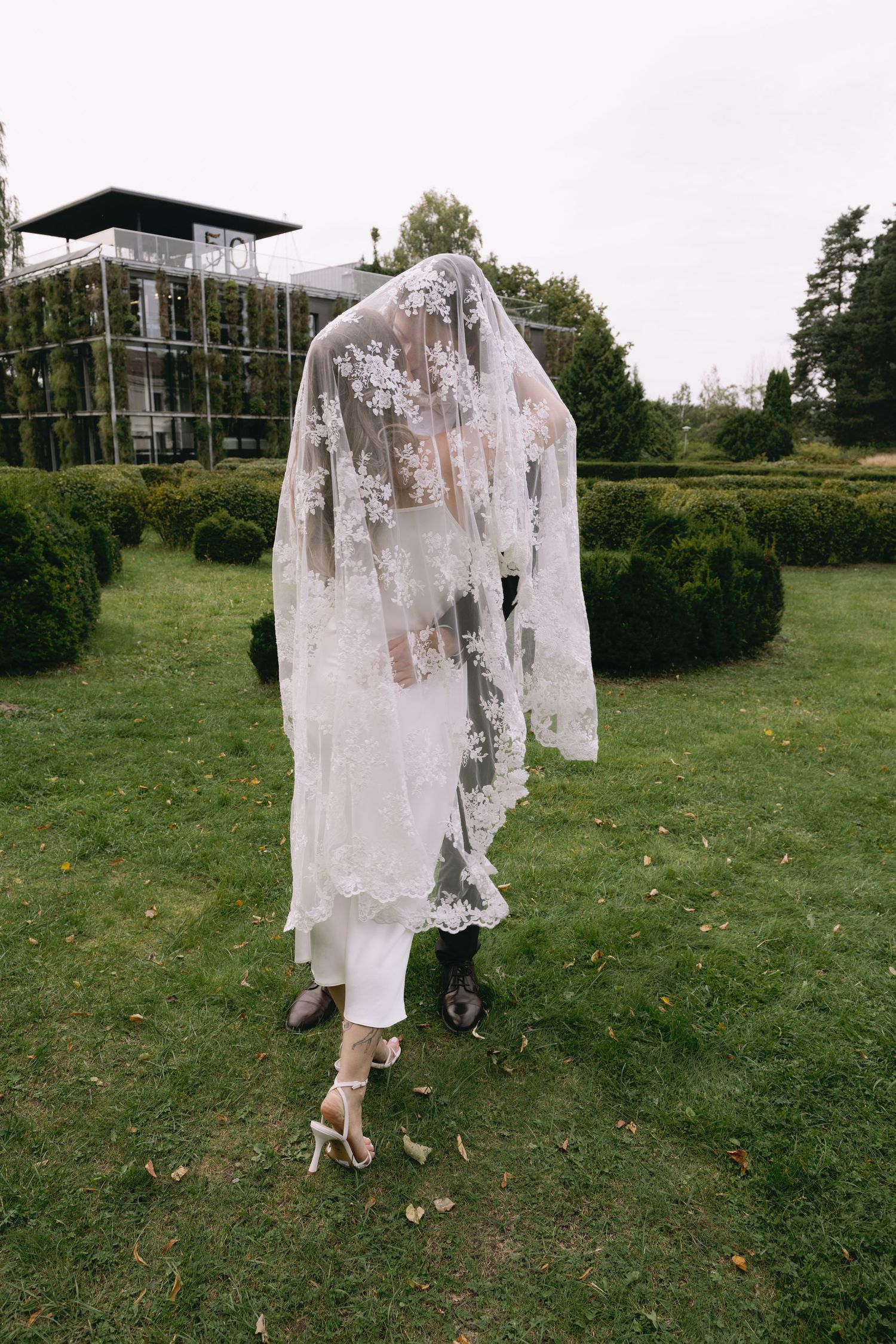 Couple walking through a manicured garden hedge maze, hidden beneath a long floral lace wedding veil, with the bride in white heels.