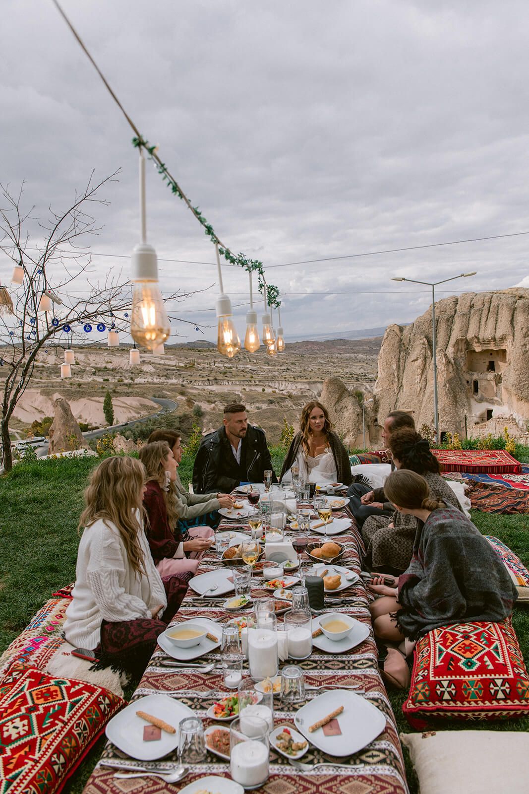 Vestuvių svečiai vakarieniauja prie ilgo stalo su tradiciniais kilimais ir girliandomis dykumoje – stilinga „destination wedding“ akimirka