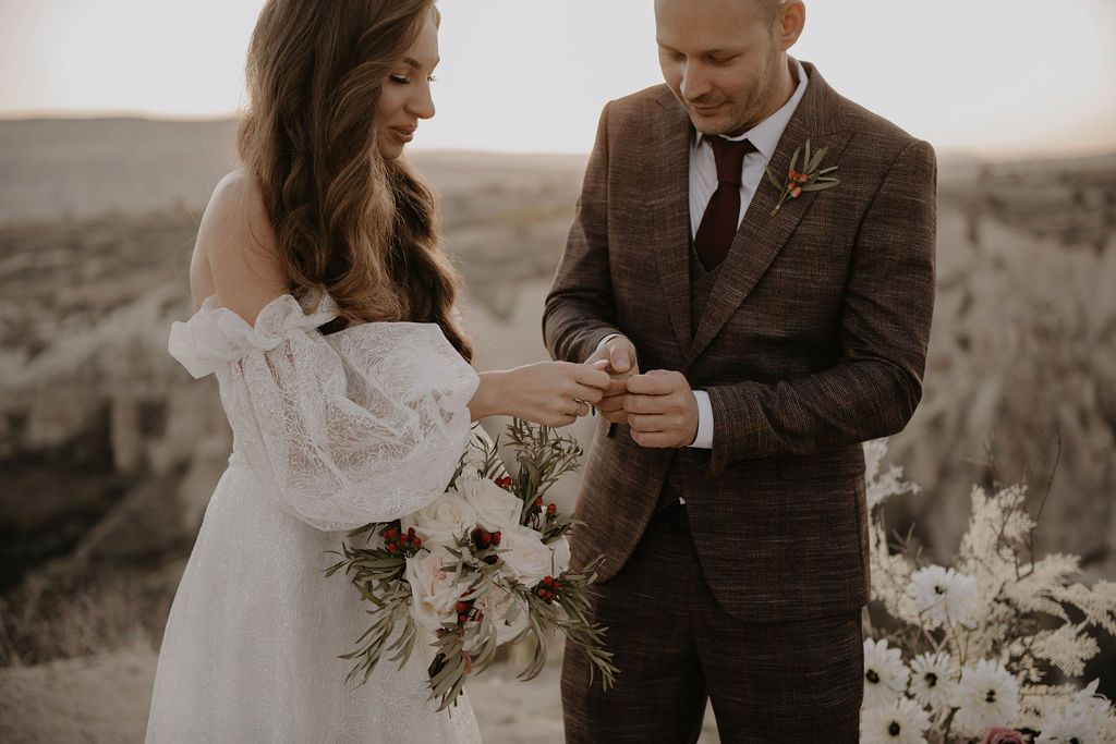 Groom placing wedding ring on bride's finger during outdoor ceremony, bride holding bouquet in lace off-shoulder gown.