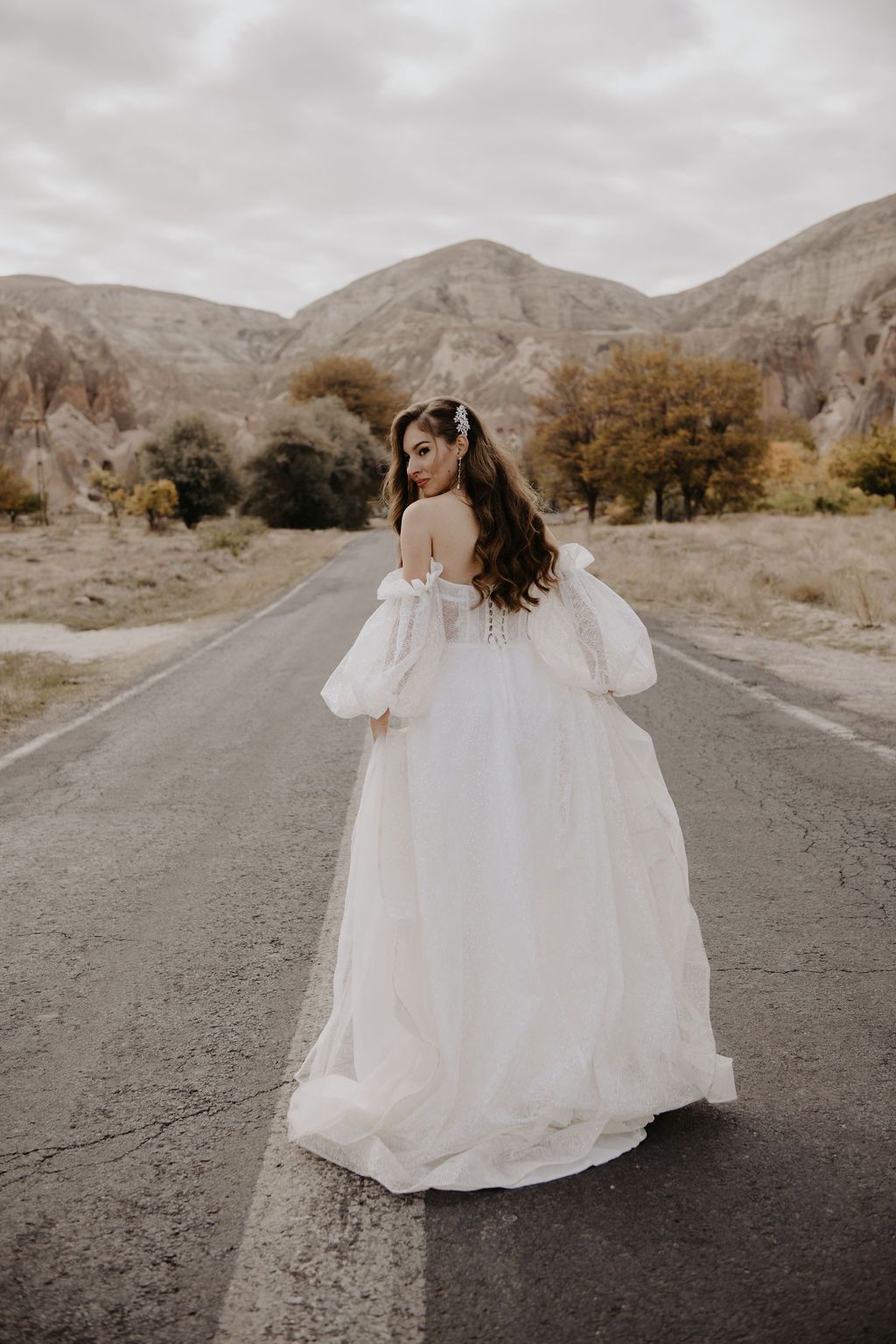Bride in a white off-shoulder ballgown with puff sleeves standing on a road with mountains behind her.