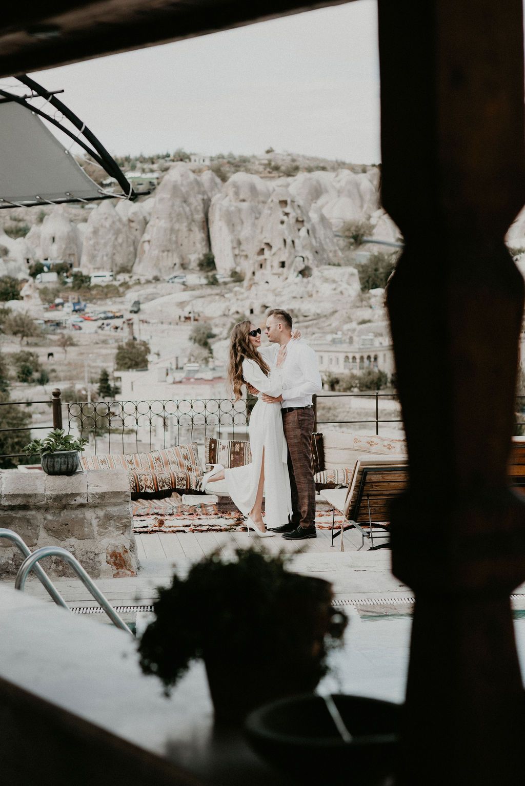 Romantic couple embracing on a terrace with stunning Cappadocia cave rock formations and cityscape visible in background.