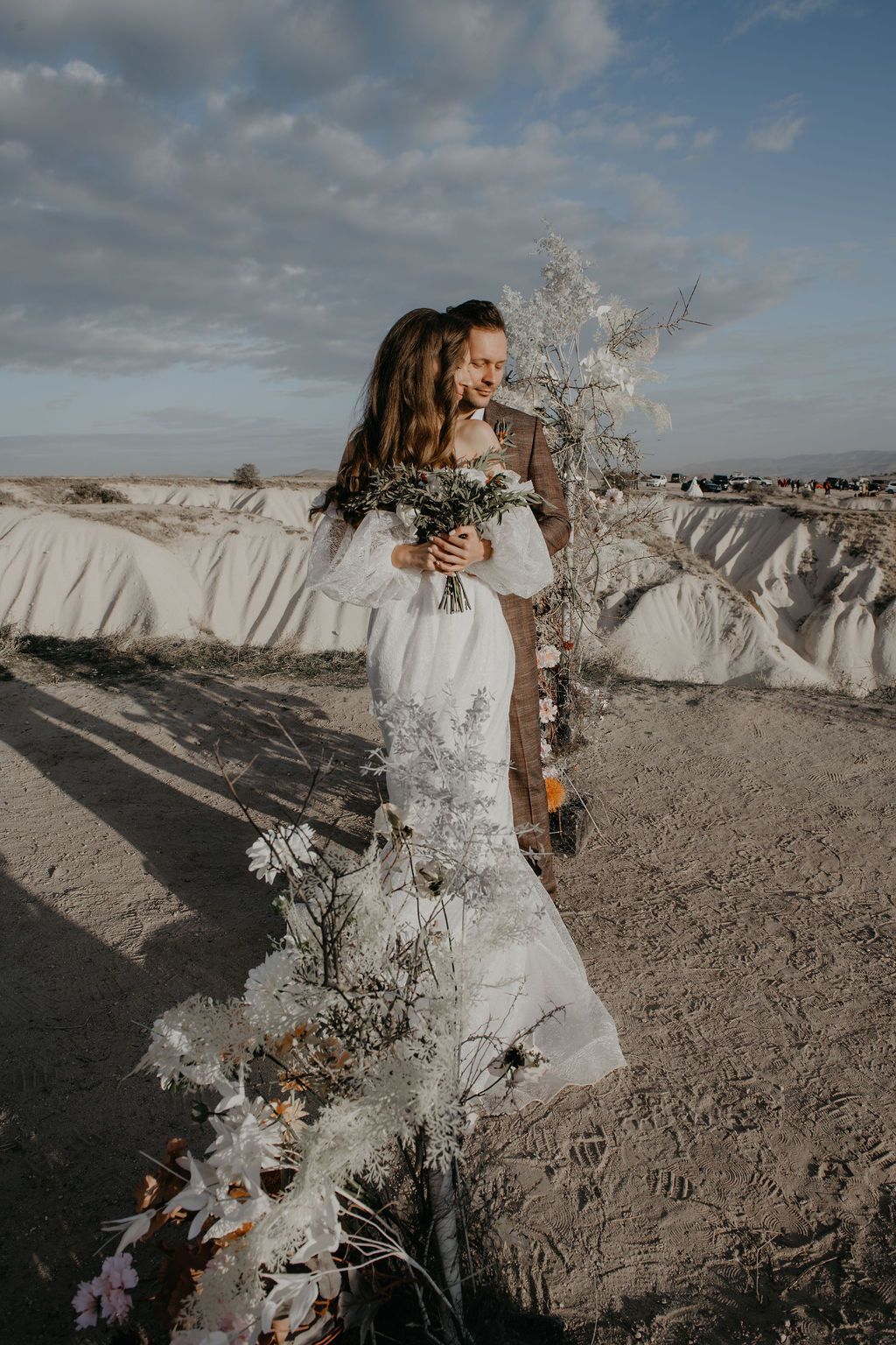 Bride in white lace gown holds bouquet as groom embraces her outdoors in sandy desert landscape with white flowers.
