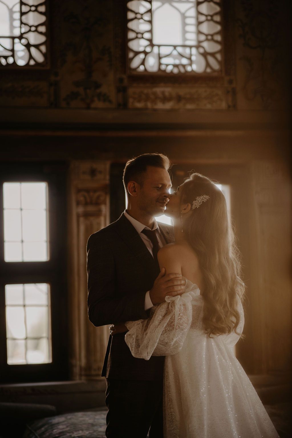 Bride and groom share romantic moment in golden sunlight inside ornate historic venue.
