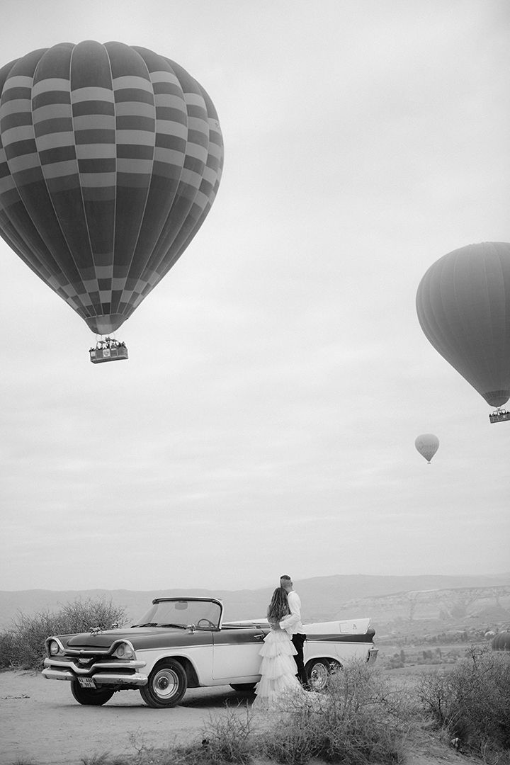 Black and white photo of a wedding couple beside a vintage convertible car with hot air balloons floating in the sky.