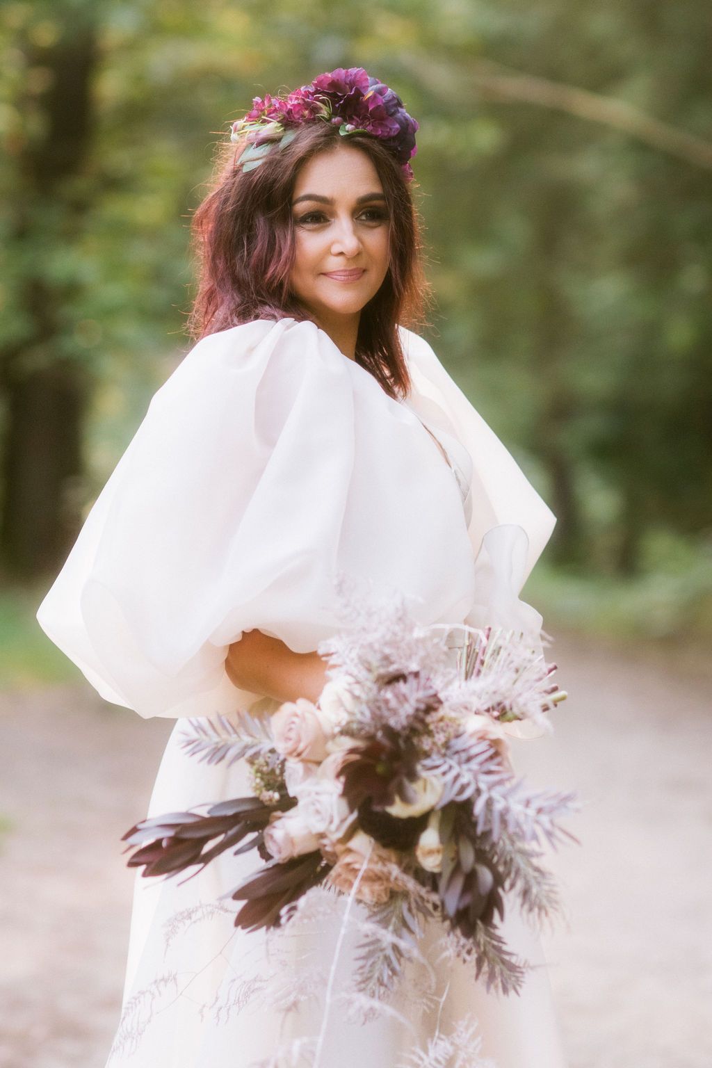 Bride in white puff-sleeve gown with floral crown holds bouquet of purple blooms in a forest setting.