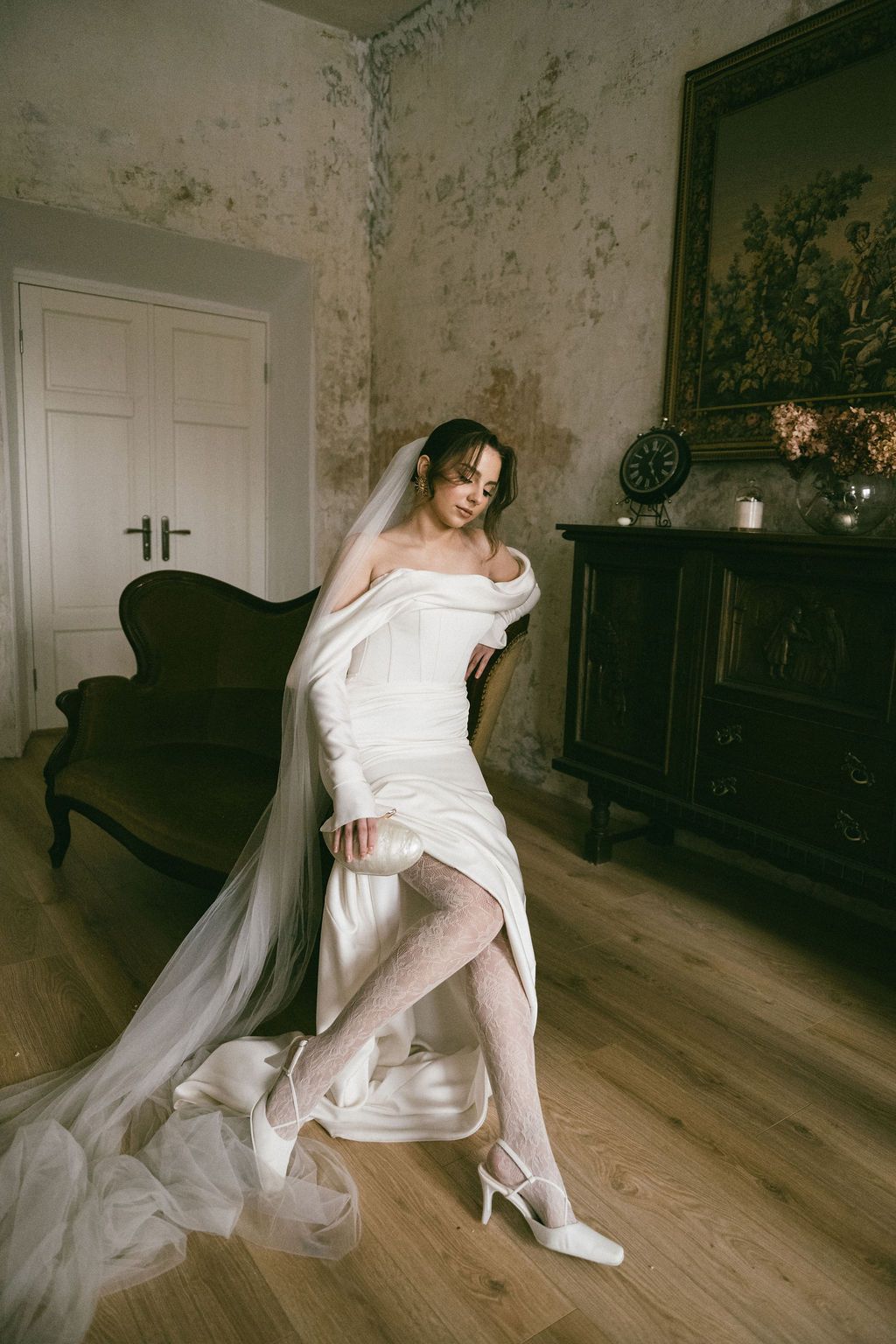 Bride in white off-shoulder gown and veil adjusting heels in vintage room with dark wood dresser.