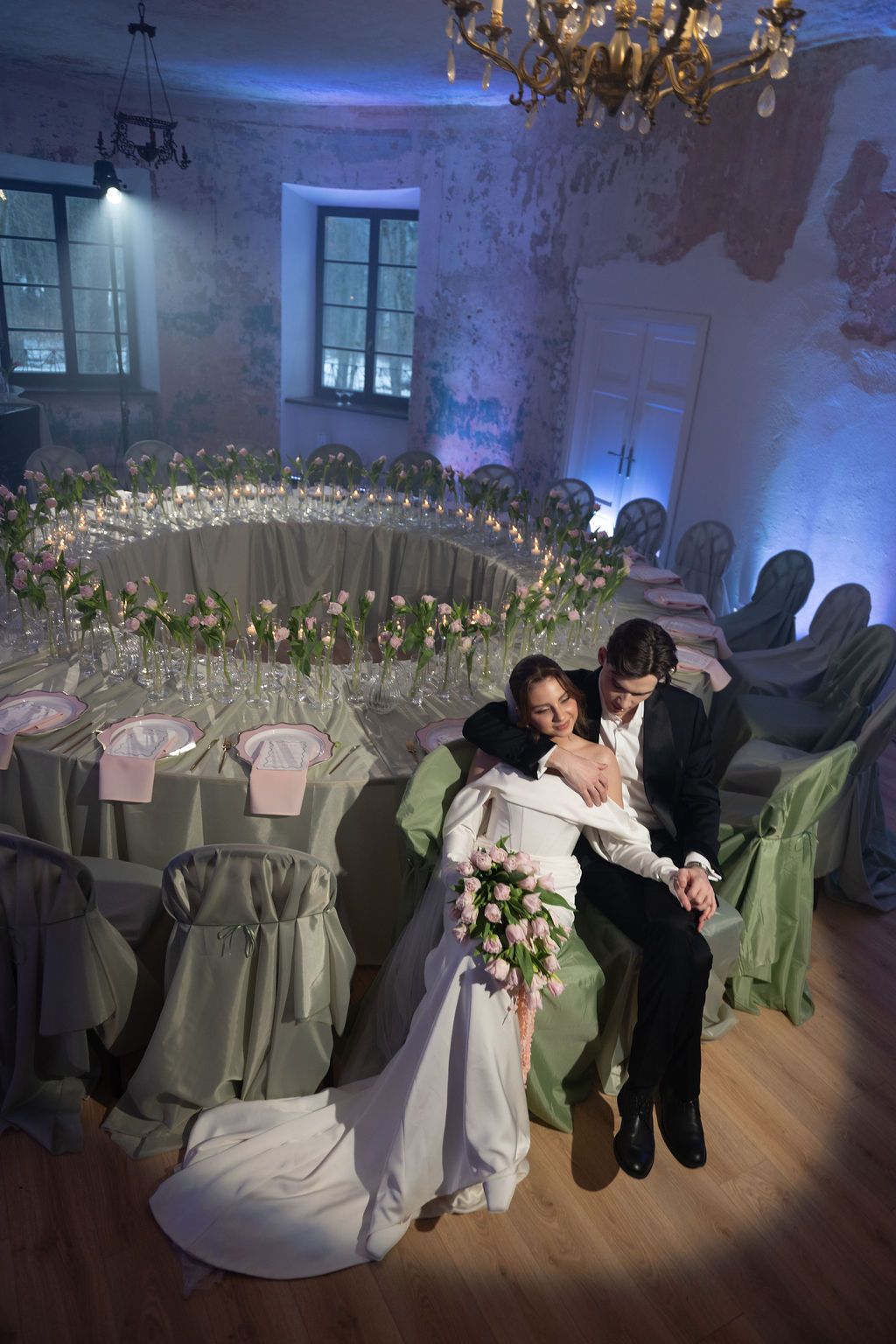 Bride and groom share an intimate moment seated in an elegantly decorated wedding reception hall with green chair covers.