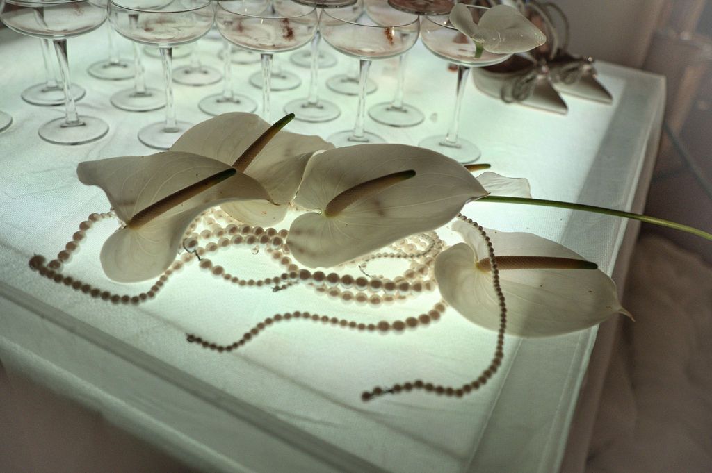 Elegant white table display with wine glasses, pearl necklace, and white calla lily flowers on a illuminated surface.