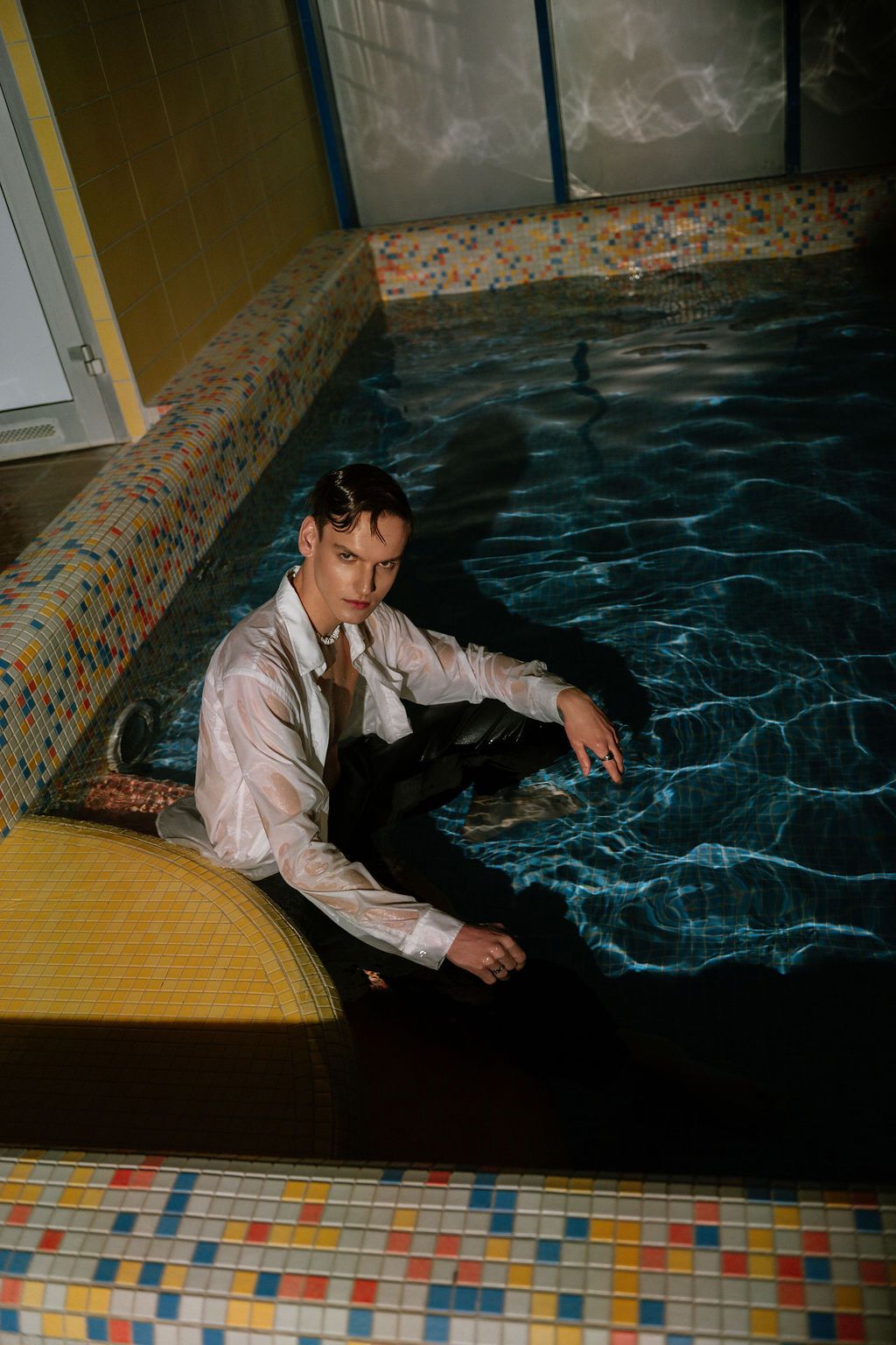 A man in a wet white shirt and dark pants sits in an indoor pool with colorful mosaic tiles.
