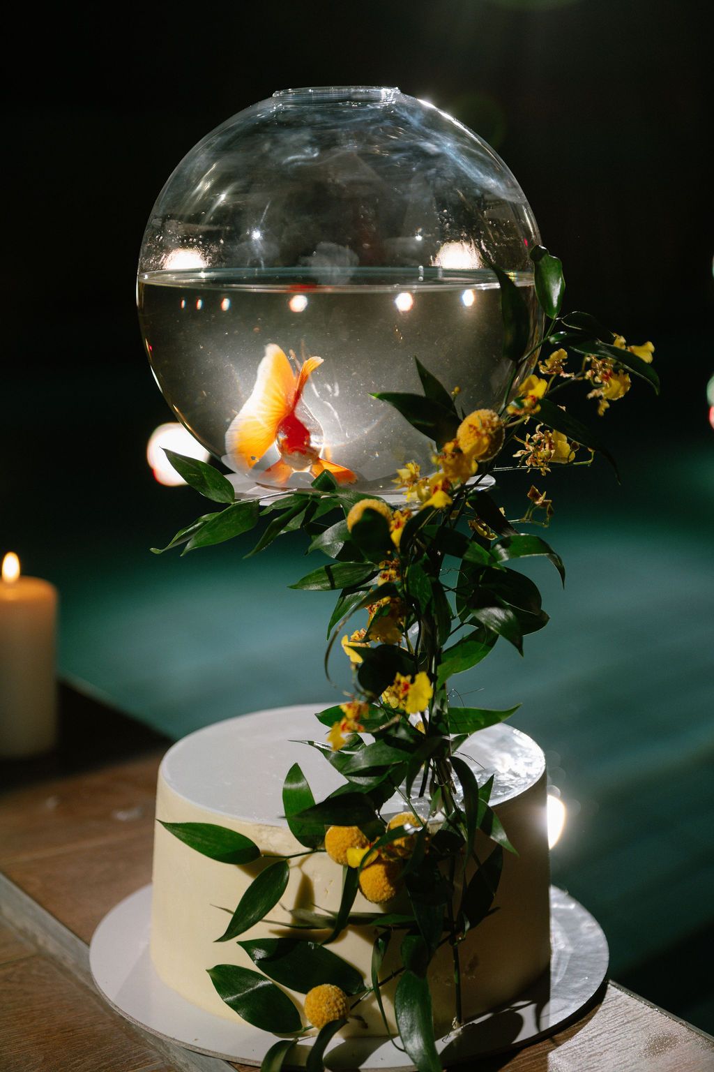 White wedding cake topped with a spherical fish bowl containing goldfish, adorned with yellow flowers and green foliage.