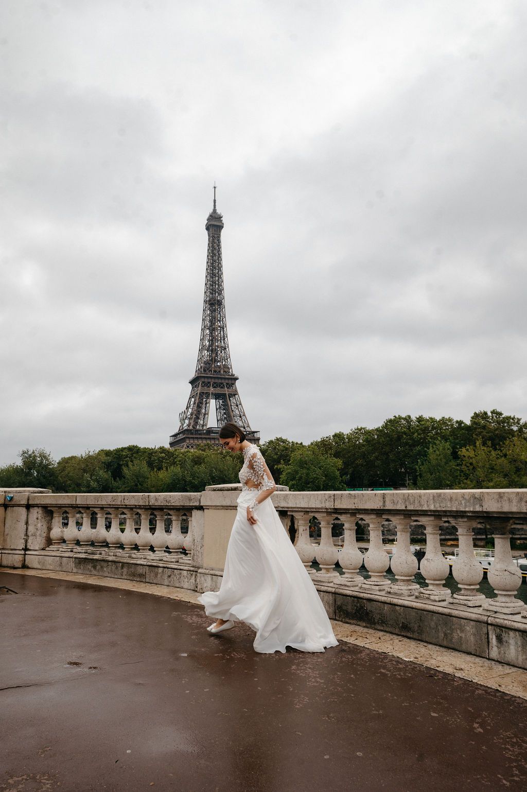 Elegant bride in white gown walking on bridge with Eiffel Tower backdrop.