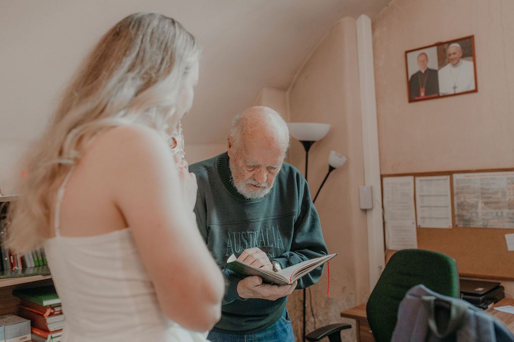 Elderly man with white beard reads a book while a young blonde woman in white top stands nearby in a cozy room.