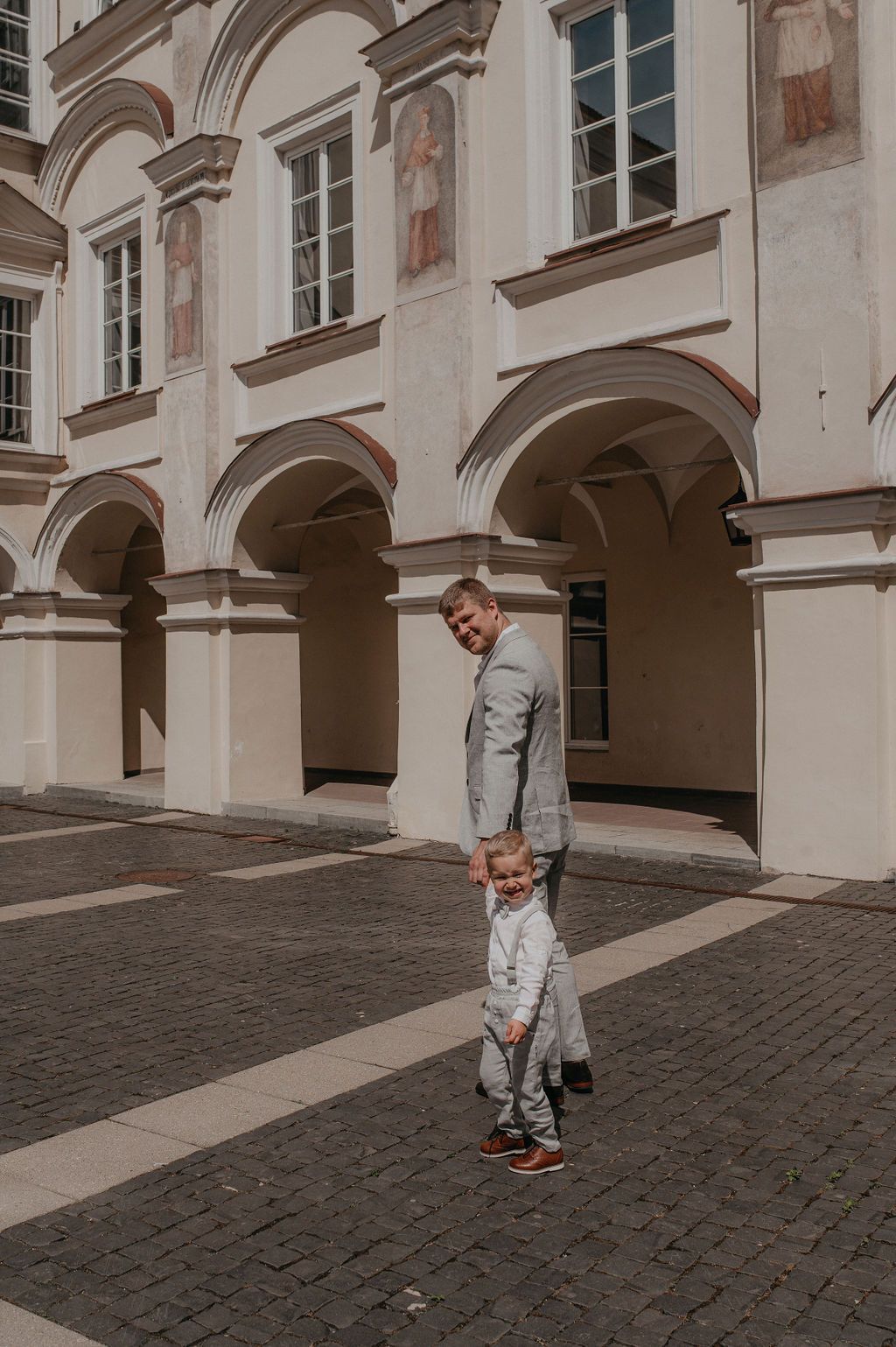 Man in gray suit holds toddler's hand in cobblestone courtyard with arched building facade.