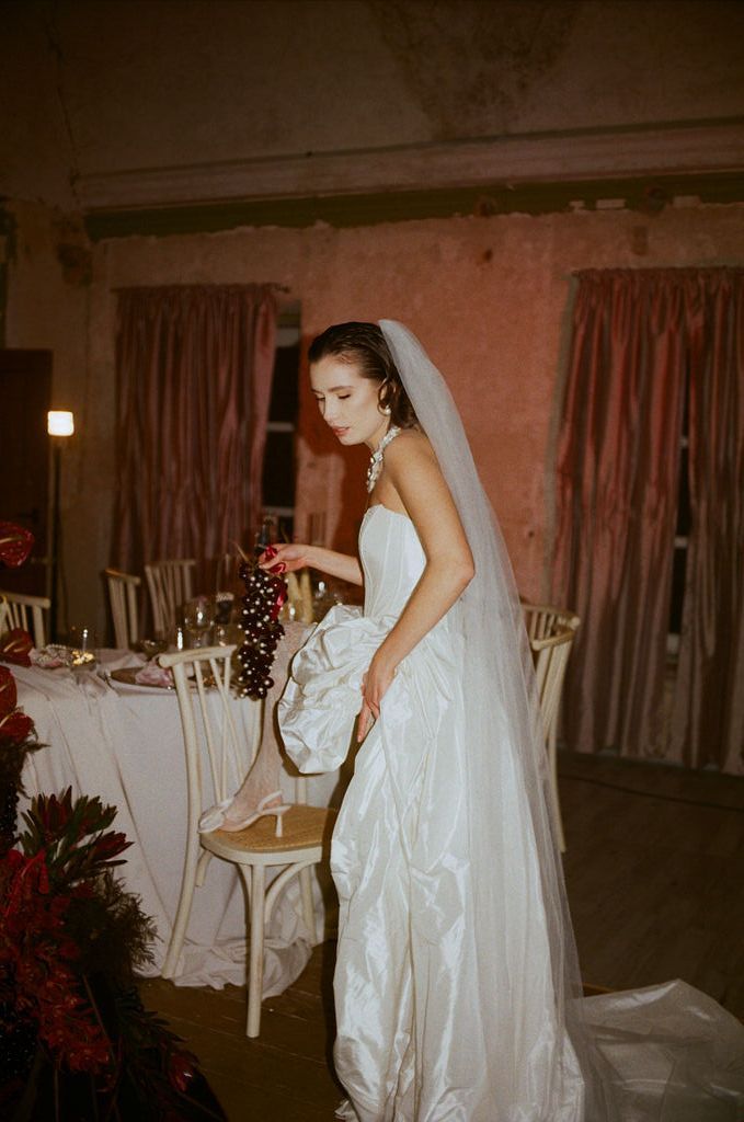 Elegant bride in strapless white ball gown with long veil stands by decorated reception table in candlelit room.