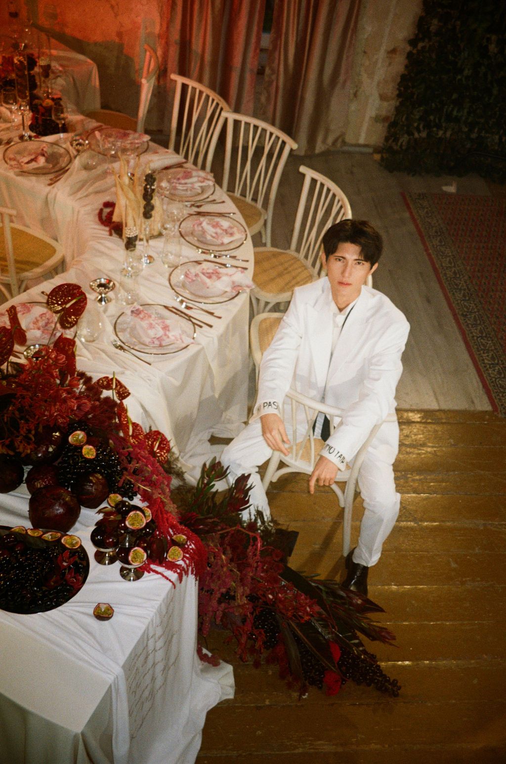 Young man in white suit seated beside an elegant red-themed banquet table with floral arrangements and fine dinnerware.