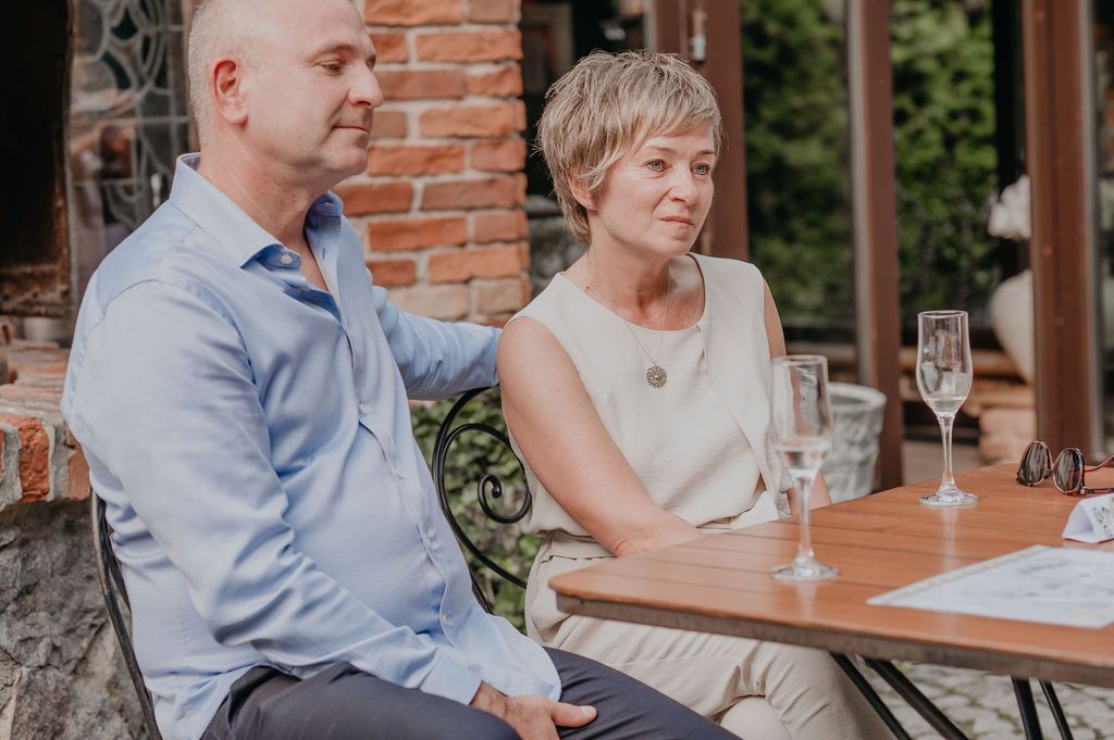 A man and woman sit together at an outdoor table with champagne glasses, dressed in smart casual attire.