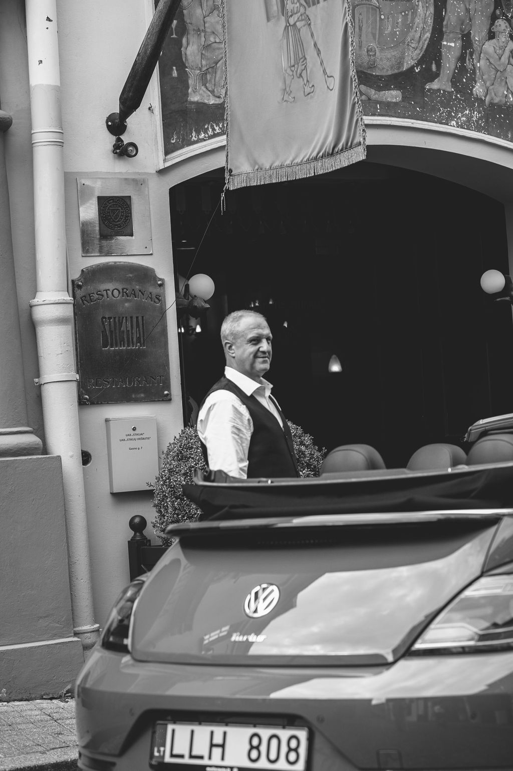 Black and white photo of elegant man in vest beside VW Beetle outside vintage restaurant entrance.