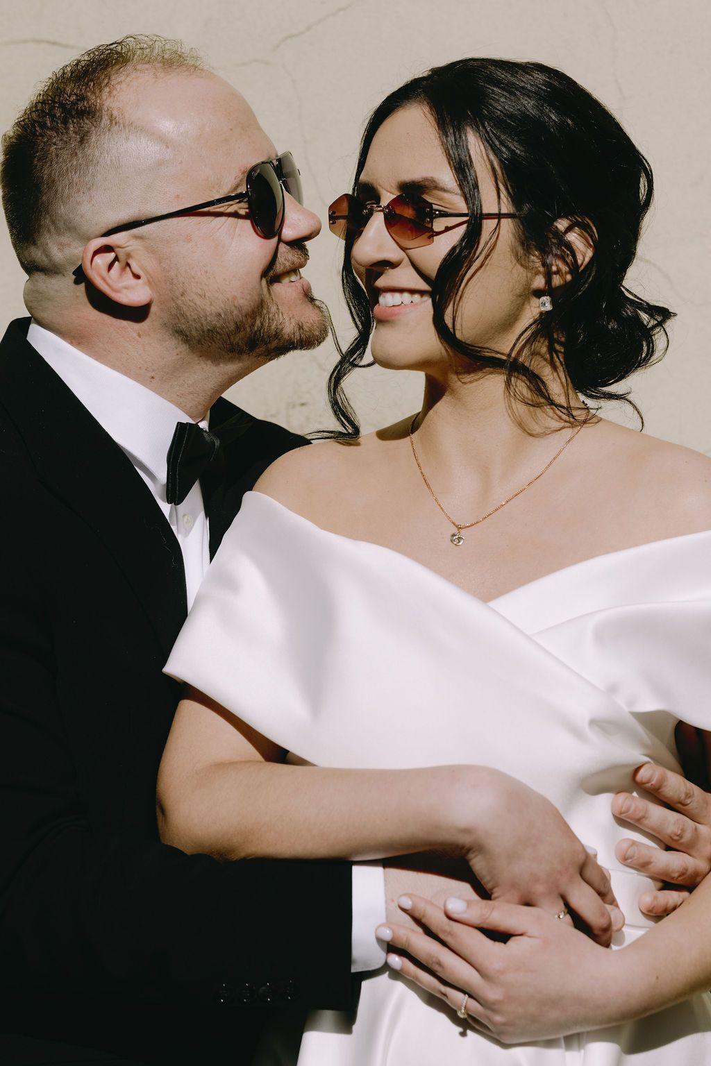 Smiling bride and groom in sunglasses share a tender moment, with groom in tuxedo embracing bride in off-shoulder gown.