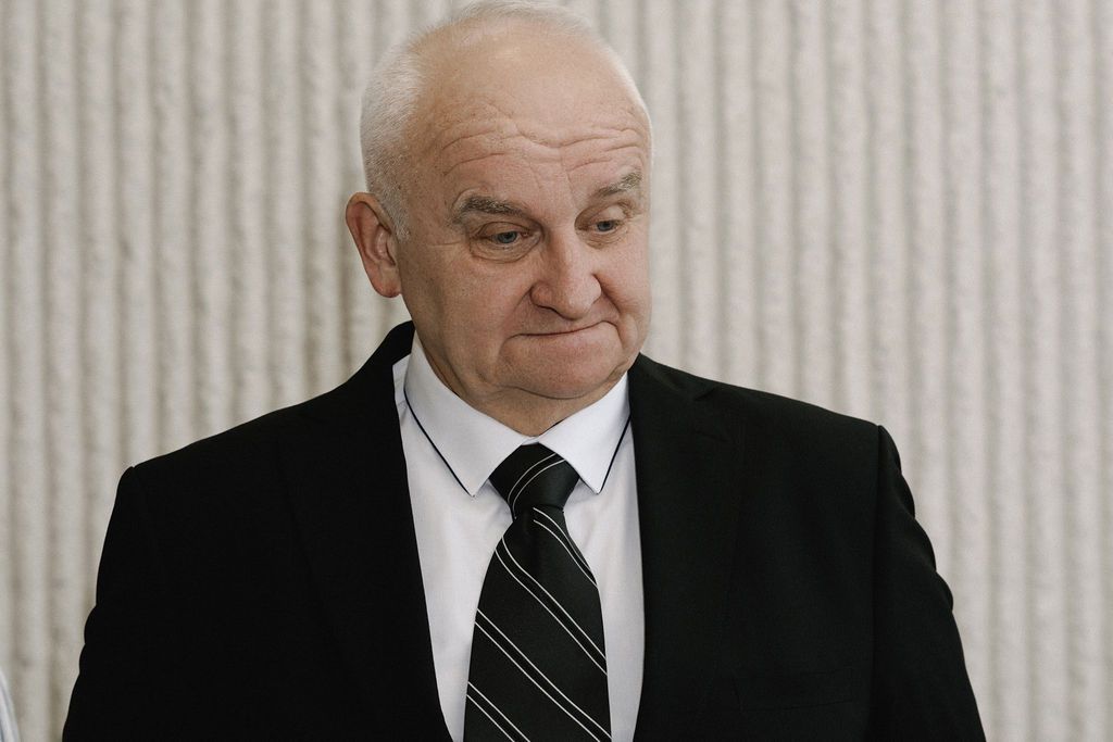 Elderly man in black suit and tie standing indoors with a serious expression.