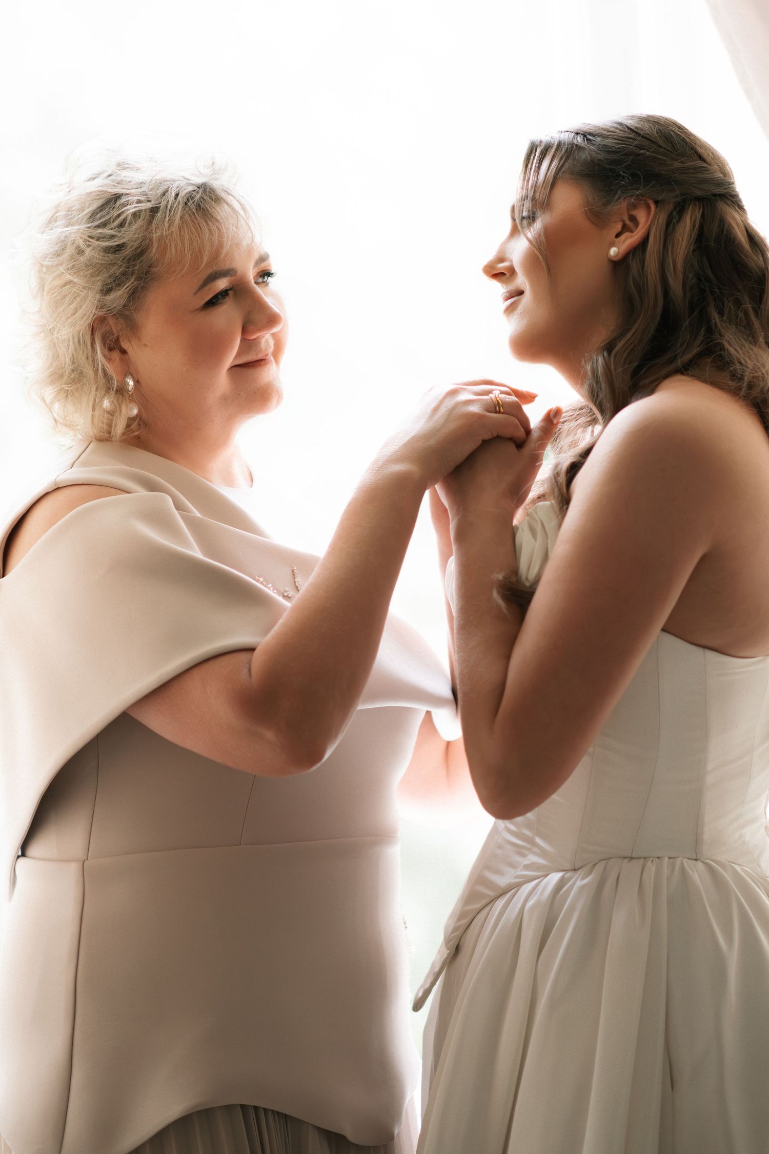 Emotional mother and bride sharing a tender moment holding hands in soft natural window light before the wedding.