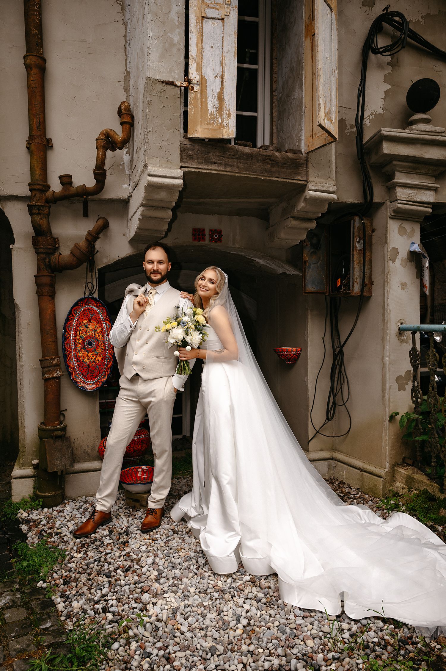 Wedding couple poses together in a rustic courtyard setting with white dress train flowing over stone ground.