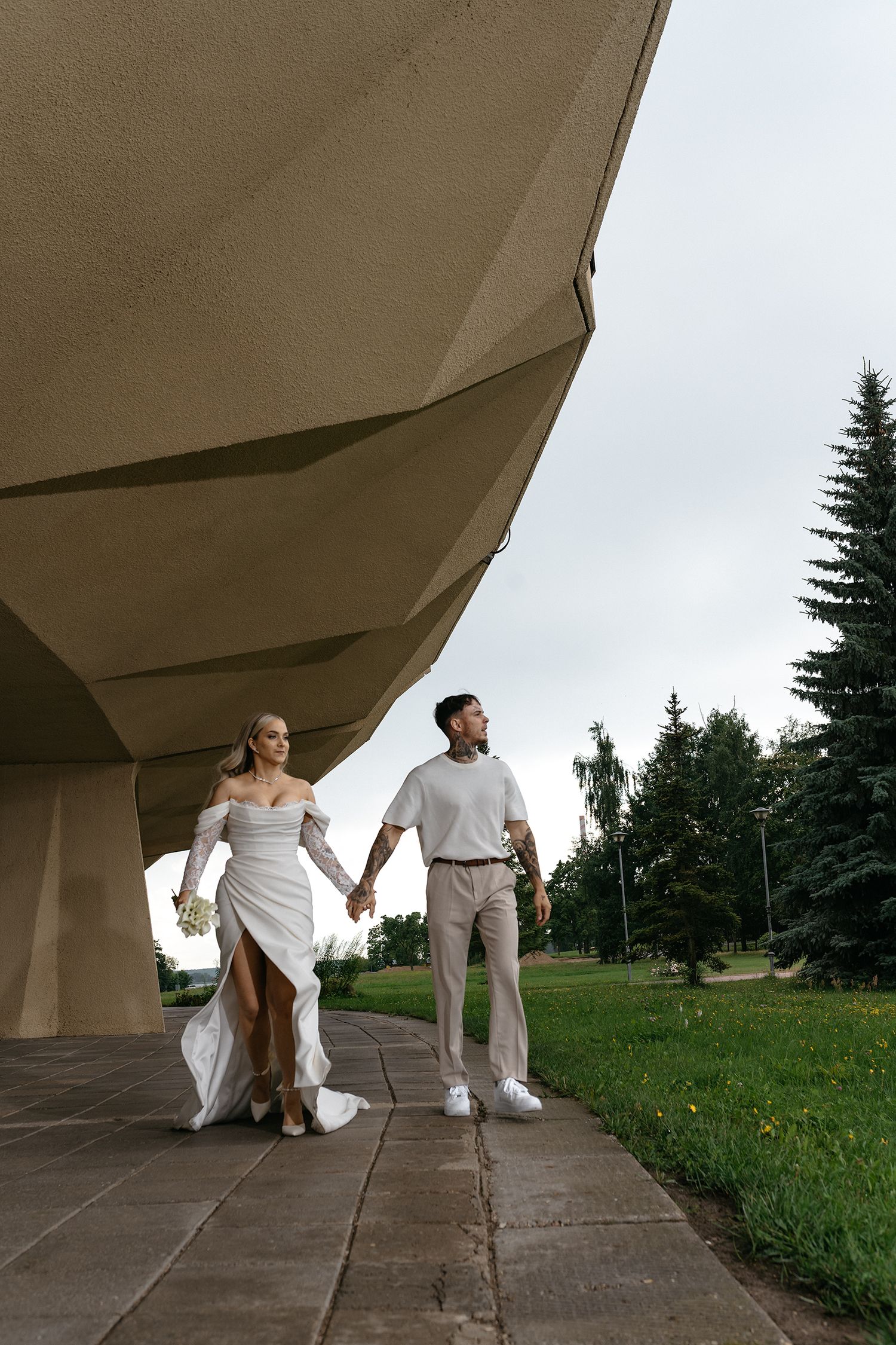 Two people in white outfits dance and pose under a concrete architectural structure in a grassy outdoor setting.