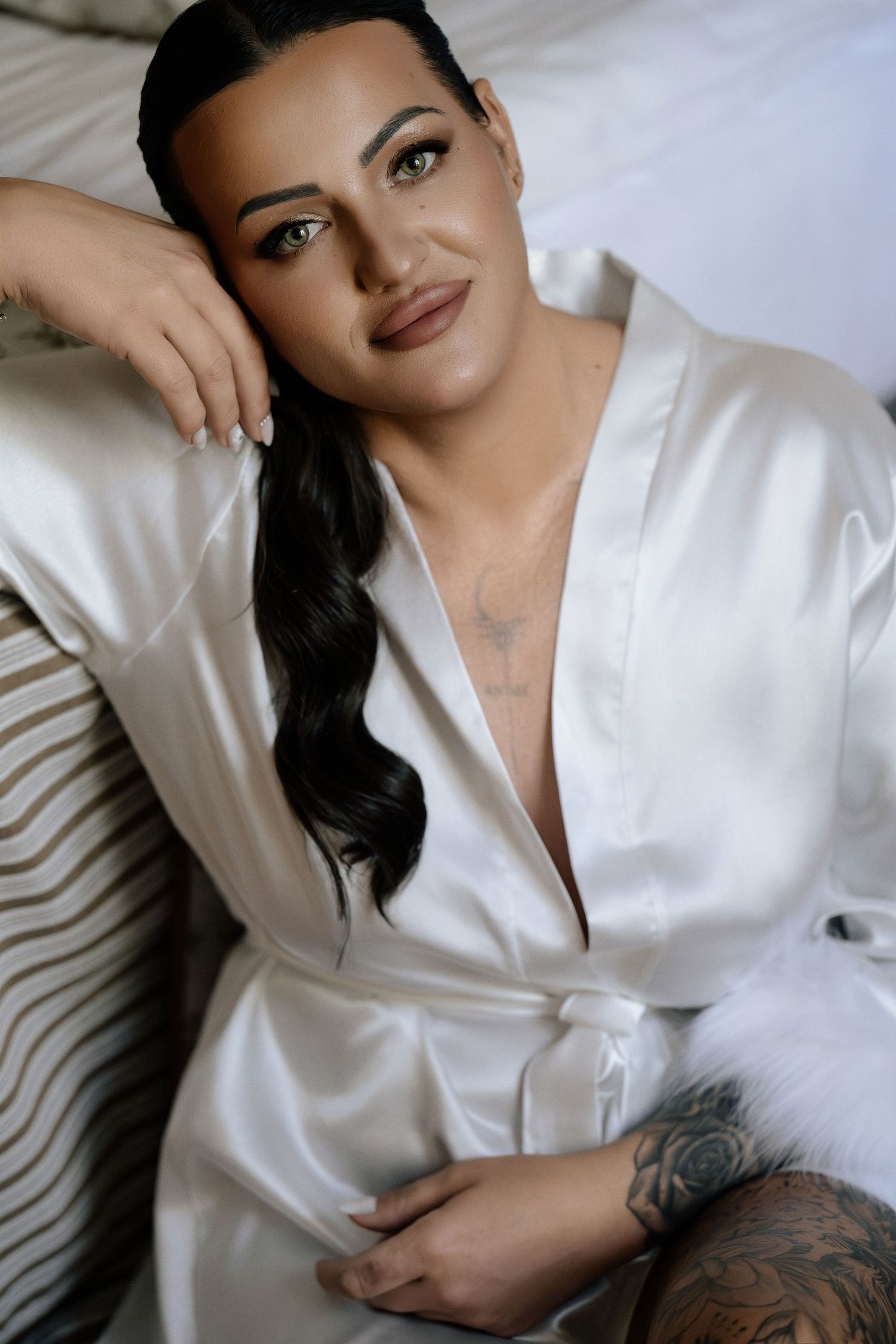 Bride with long wavy dark hair relaxes in a white satin robe, smiling softly during her wedding morning preparations.