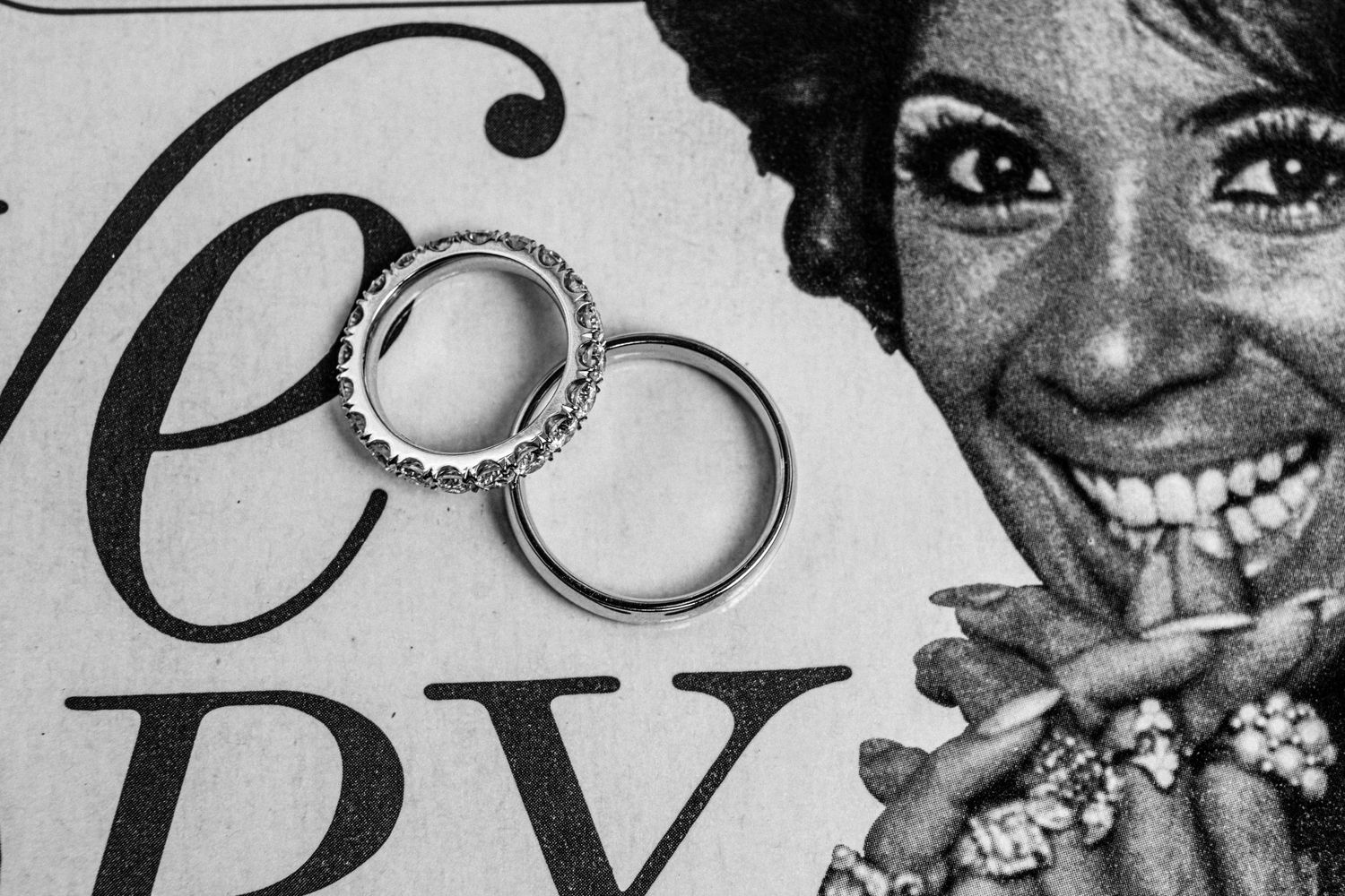 Black and white photo of two wedding rings resting on a vintage magazine page showing a smiling woman wearing a jeweled cocktail ring.