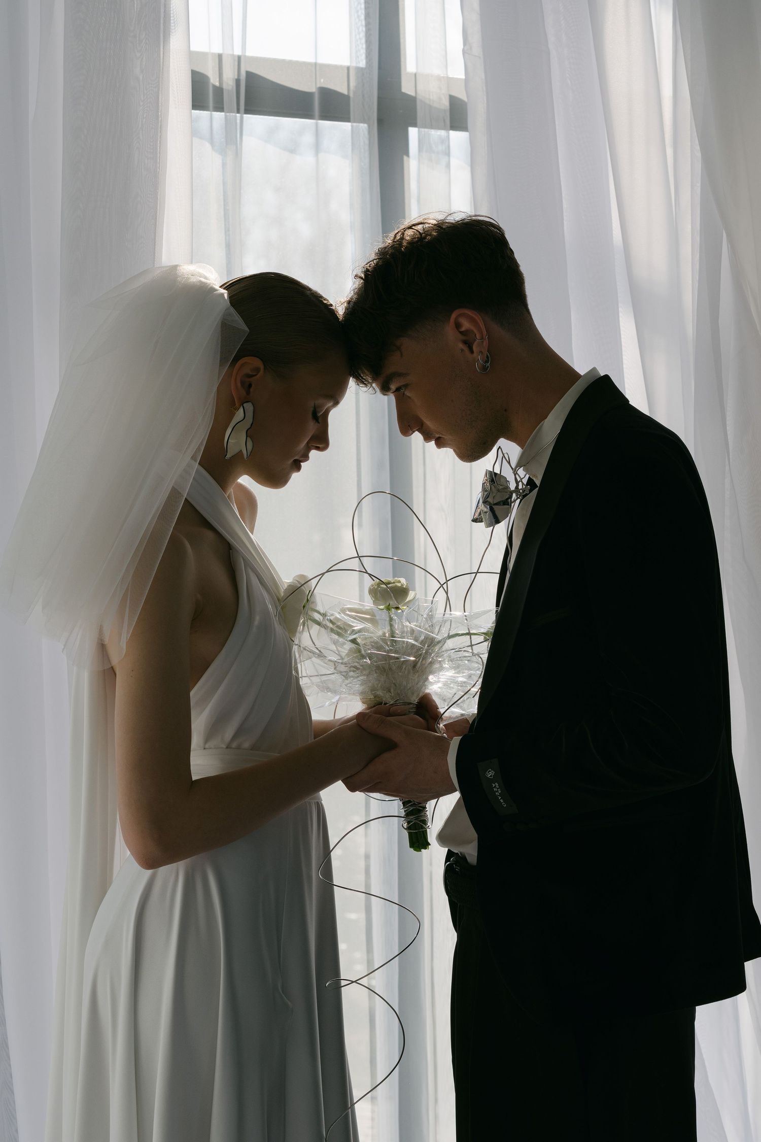 A silhouetted bride and groom share an intimate moment by a window with sheer white curtains.